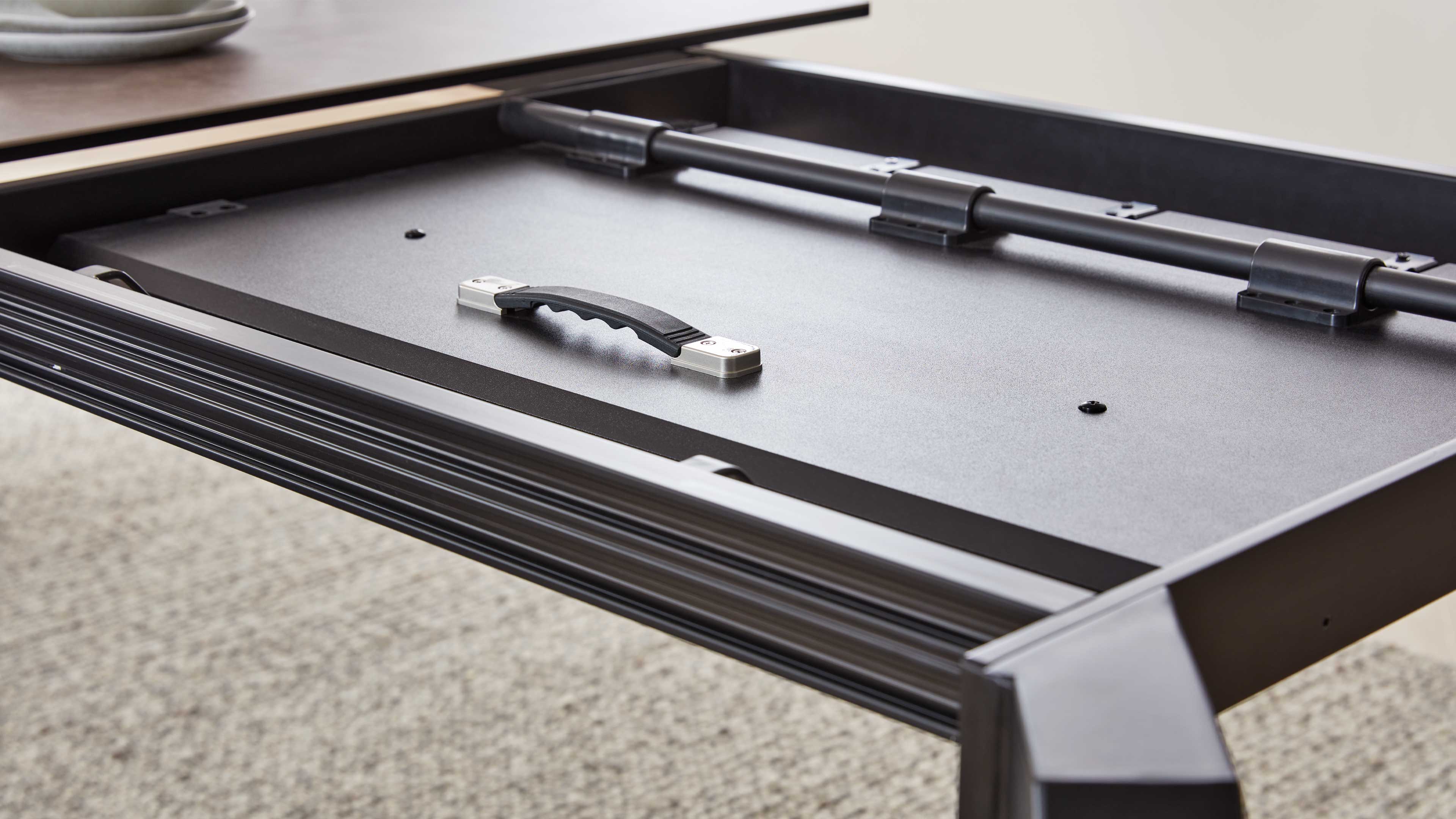 Unterseite einer ausziehbaren Tischplatte mit schwarzem Kunststoffgriff auf Metallbefestigungen und einer Rohrstrebe.