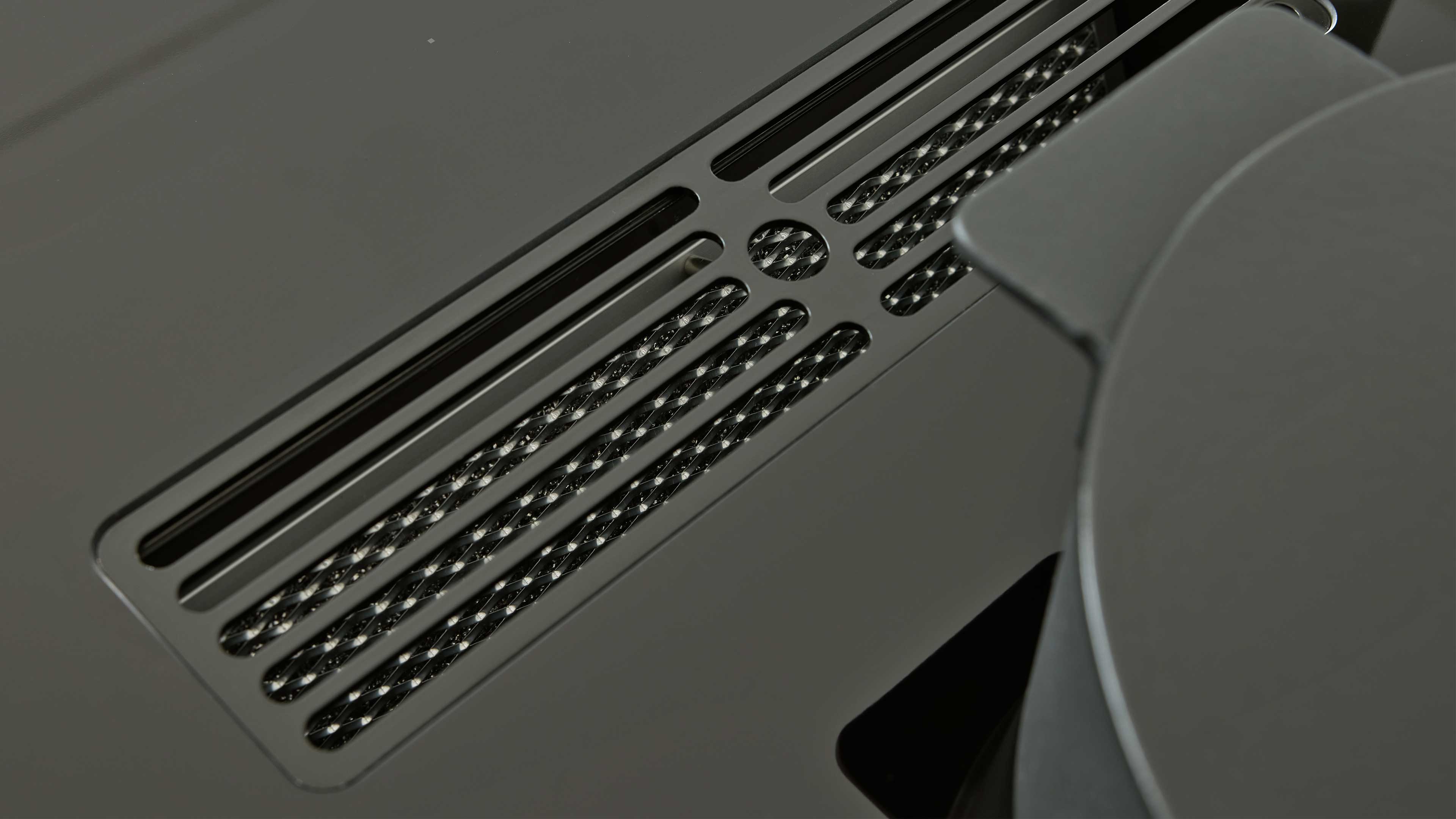 Schwarze Metall-Entlüftungsblende mit länglichen Schlitzen und sichtbarem gekreuztem Drahtgeflecht, rechts Teil einer runden matten Fläche.