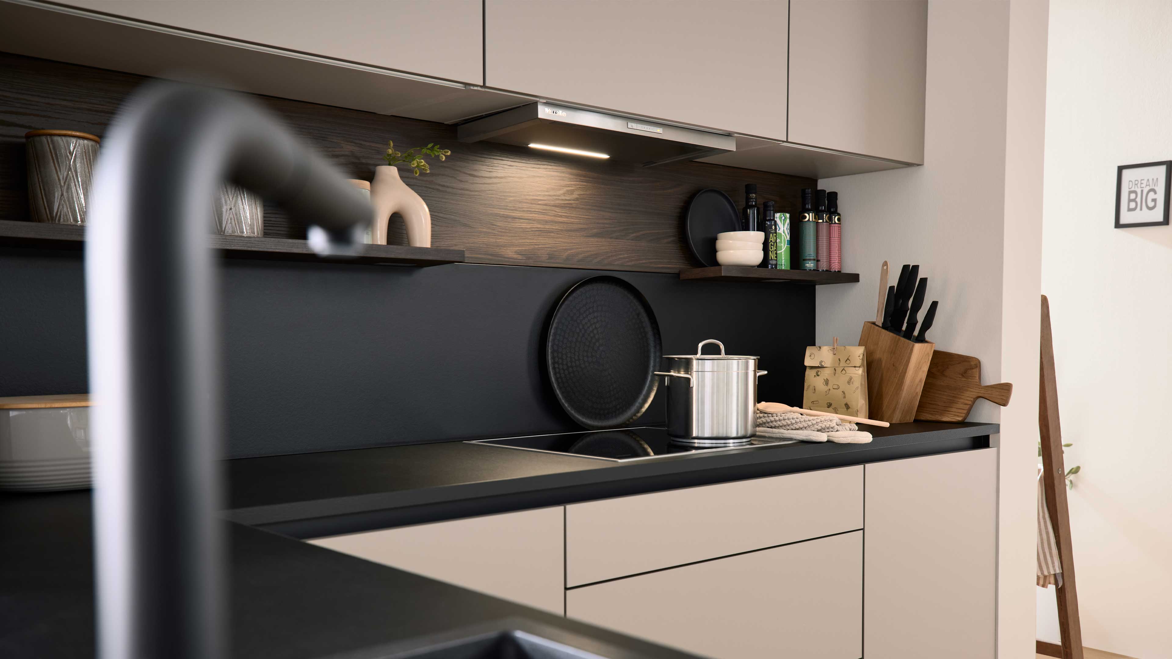 Schwarze Arbeitsplatte mit Induktionsfeld und Edelstahlkochtopf vor dunkler Rückwand, darüber Regale, Messerblock und Holzbrett.