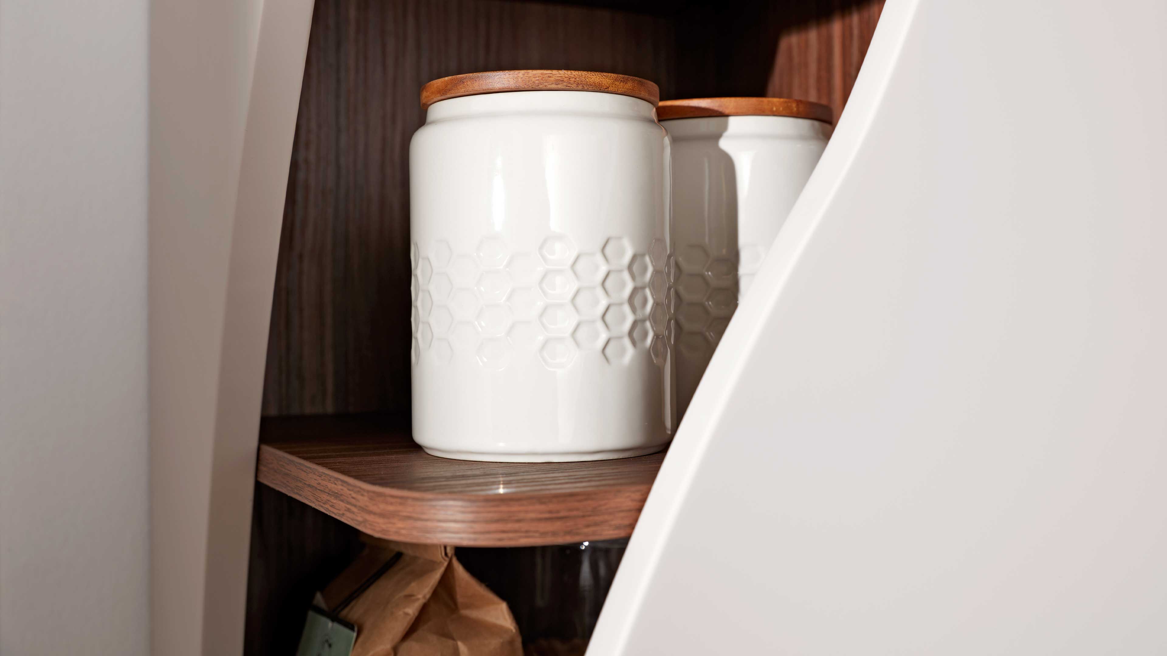 Zwei weiße, glänzende Vorratsdosen mit Wabenprägung und runden Holzdeckeln stehen auf braunem Regalbrett in einem weißen Schrank.