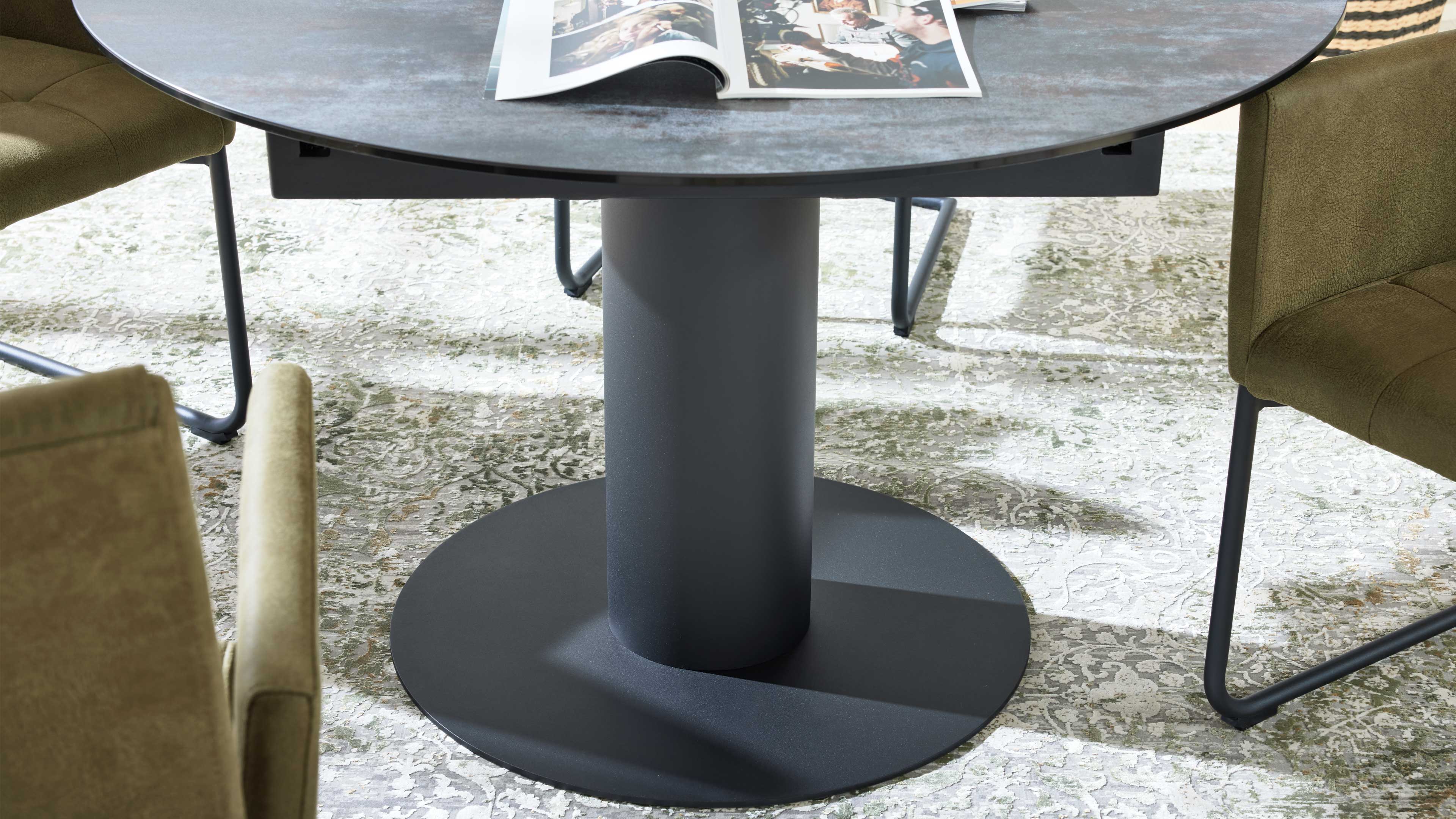 Runder Tisch mit dunkelgrauem, zylindrischem Fuß auf runder Bodenplatte, offenes Magazin oben, grüne Stühle auf gemustertem Teppich.