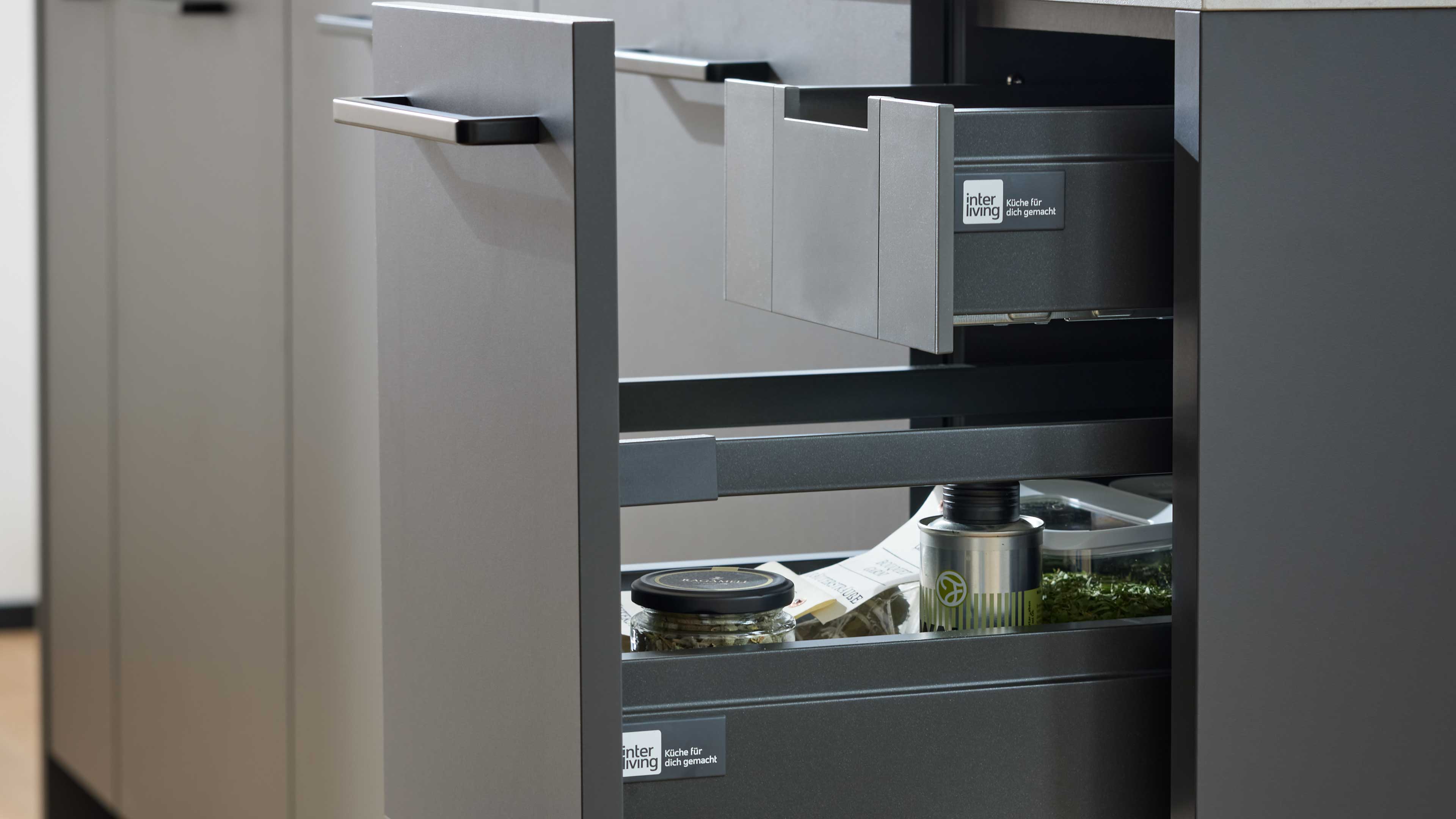 Geöffnete graue Küchenschubladen mit flachen Metallgriffen, darin ein Glas, eine Dose und lose Kräuter.