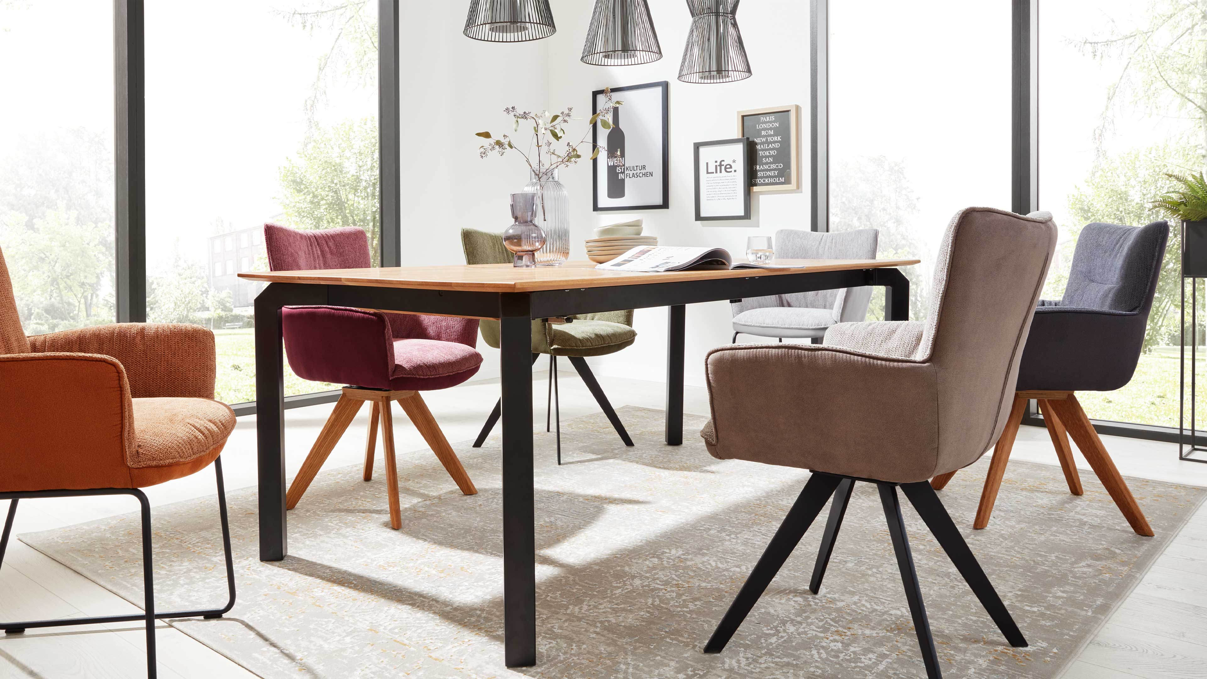 Rechteckiger Holztisch mit schwarzen Metallbeinen, umgeben von sechs verschiedenen gepolsterten Stühlen vor großen Fenstern.
