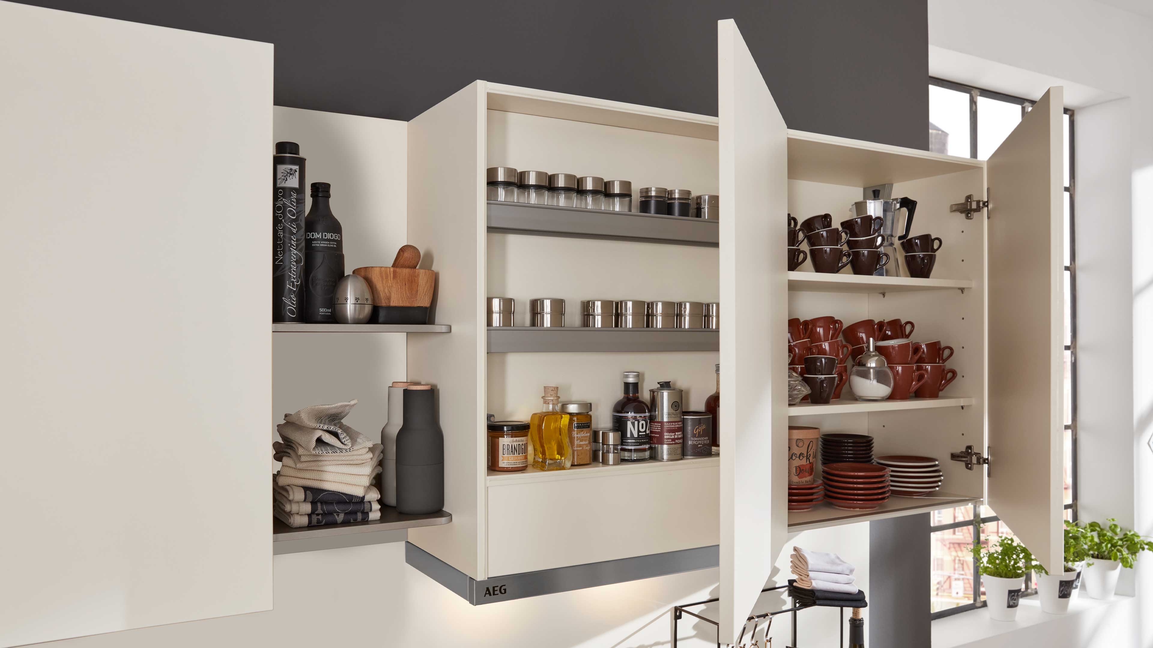 Offene, matte cremefarbene Küchenhängeschränke mit Einlegeböden, Gewürzdosen, Ölflaschen, gestapelten braunen Tassen, Tellern und gefalteten Tüchern.