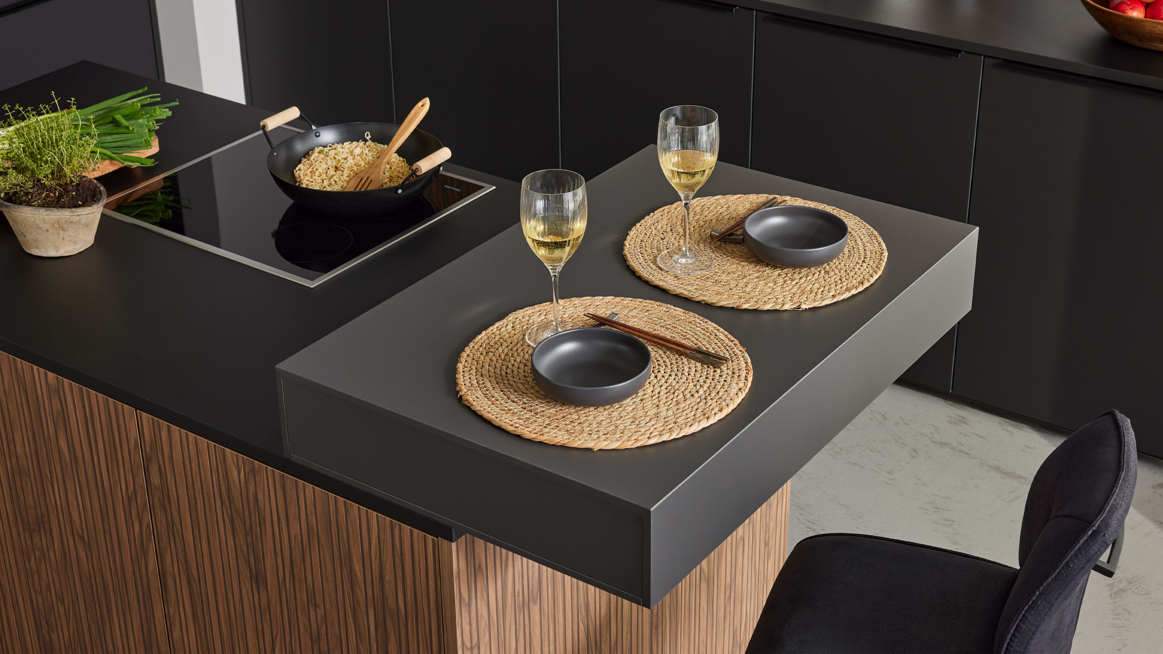 Auskragende Kücheninsel mit zwei Platzsets aus Flechtmatten, grauen Schalen, Stäbchen und gefüllten Weingläsern.