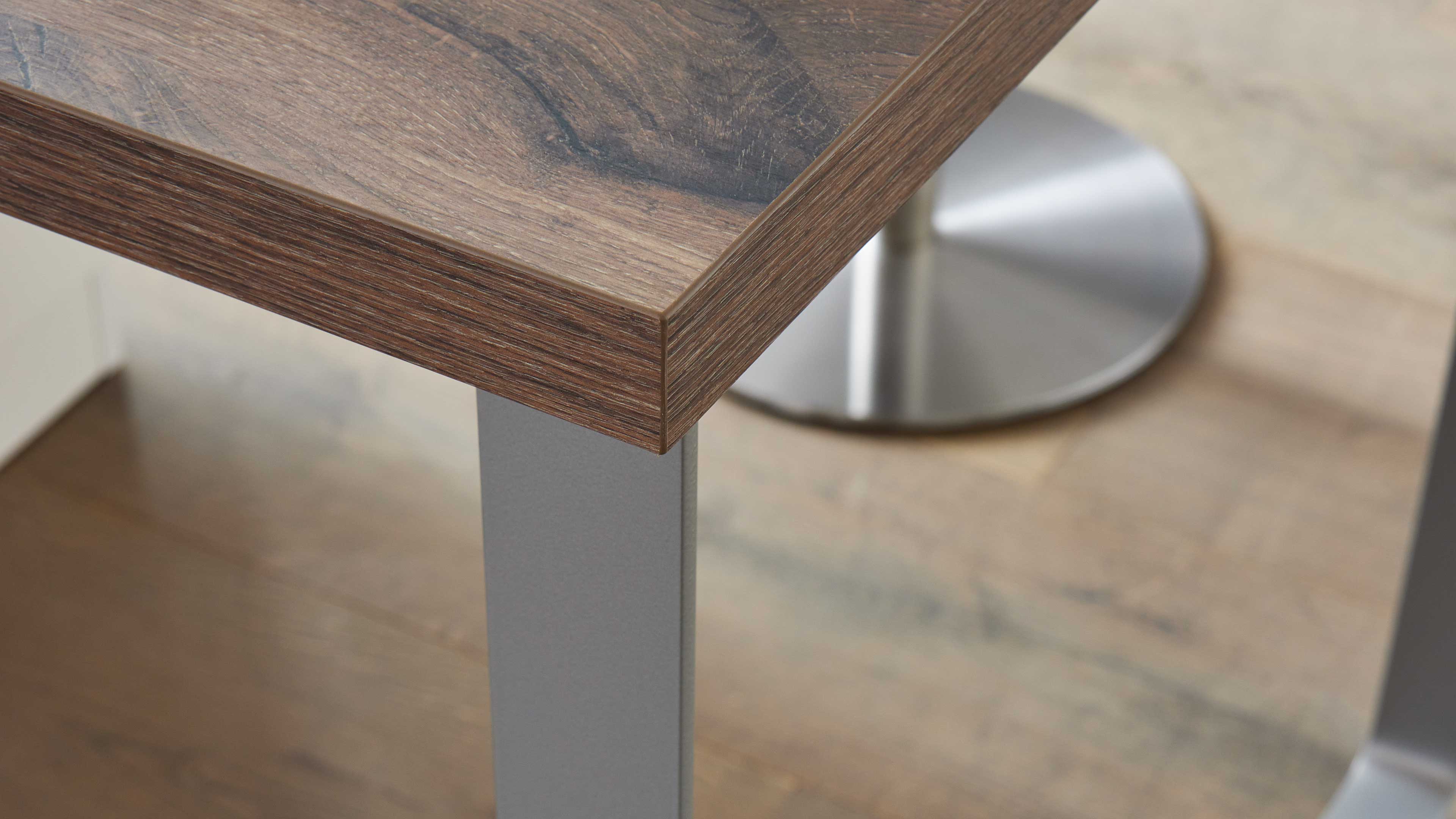 Tischplatzecke mit dunkelbrauner Holzmaserung und geschlossener Kante, sichtbares graues Metallbein und runder Edelstahlfuß auf Holzboden.