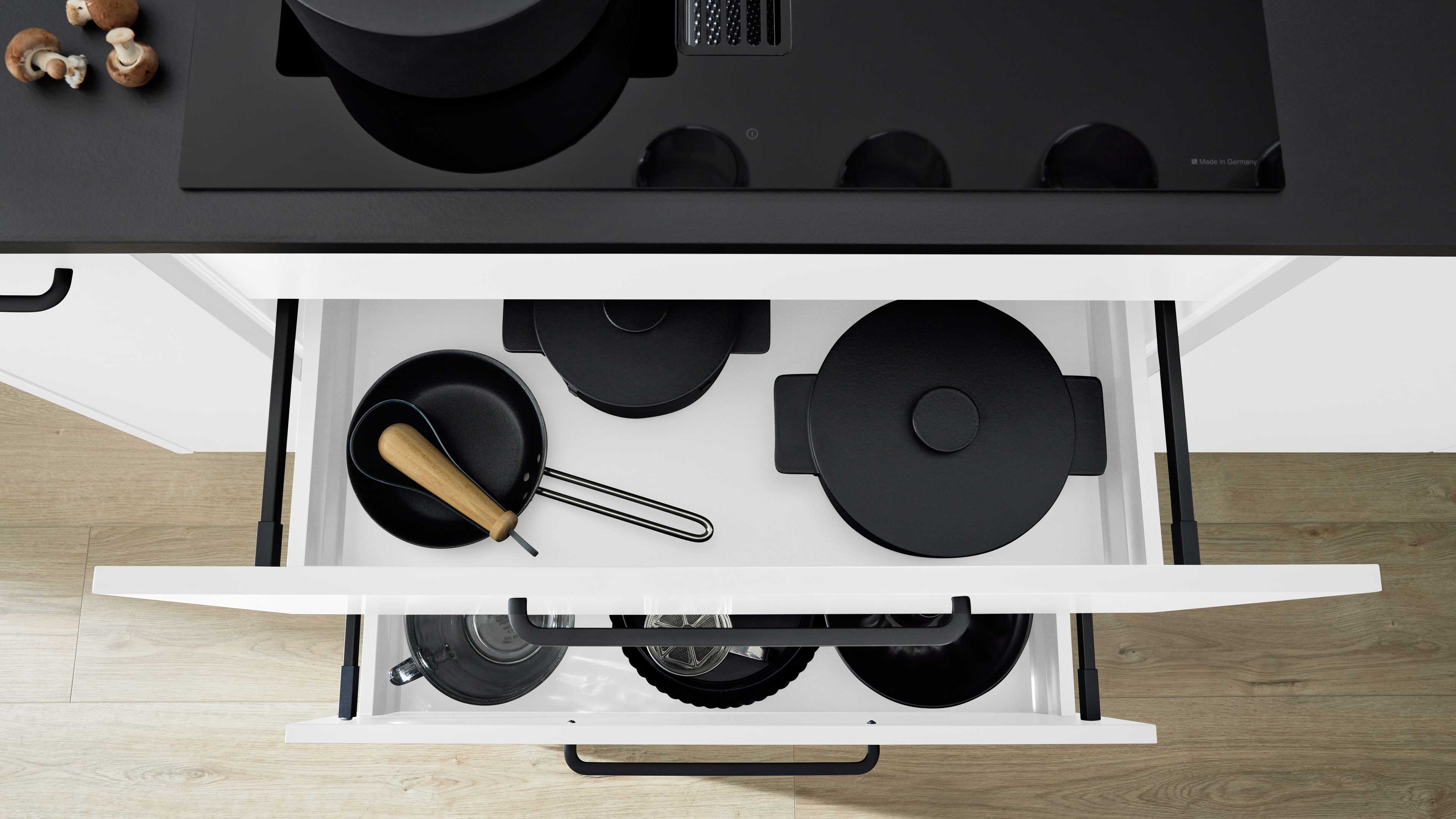 Offene weiße Küchenschublade unter schwarzem Kochfeld mit schwarzen Töpfen und Pfannen, eine kleine Pfanne mit Holzgriff.