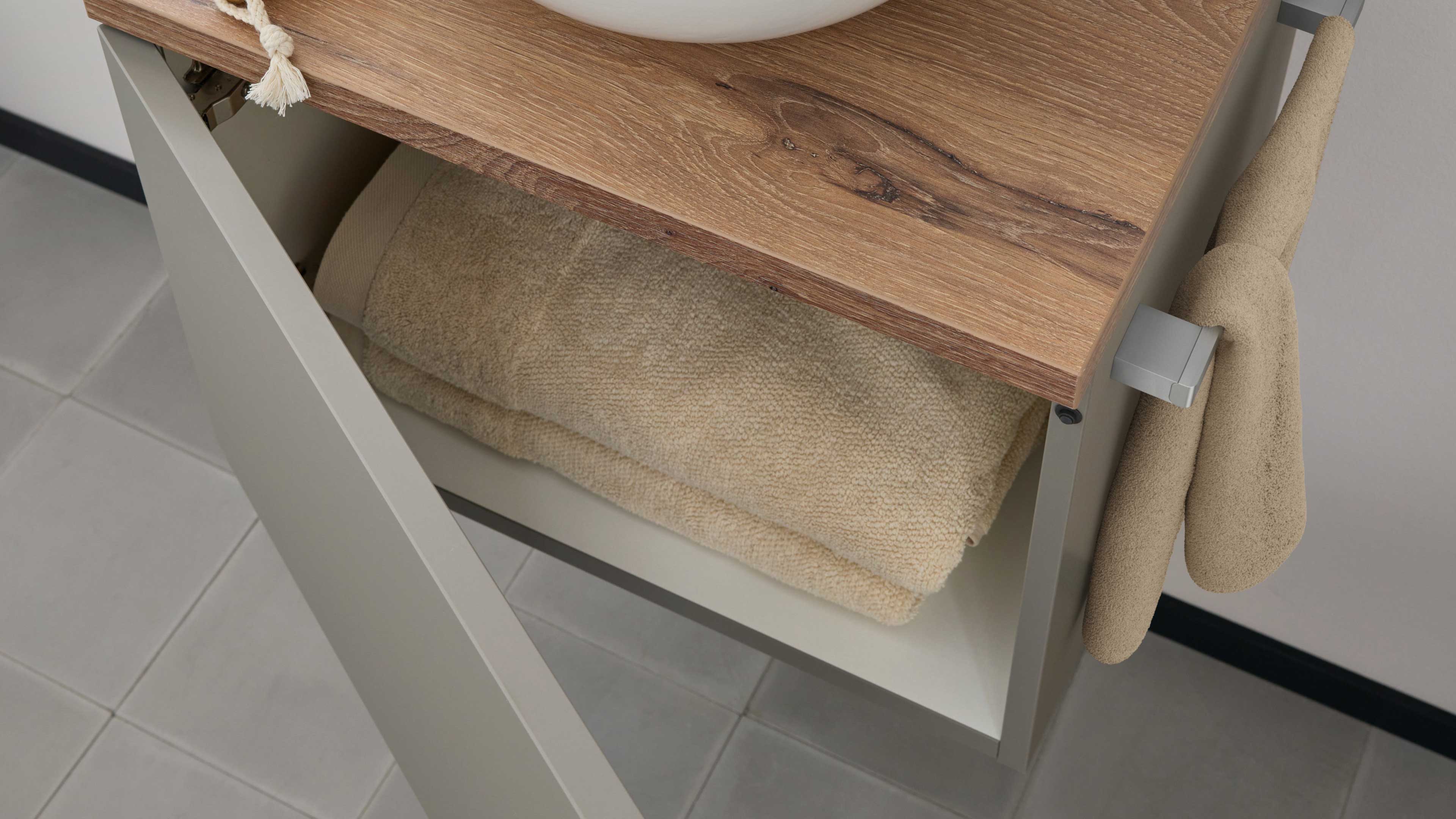 Offenes graues Unterschrankfach mit hellen, gefalteten Handtüchern unter einer Holzplatte, seitlich ein Haken mit beigen Handtüchern.