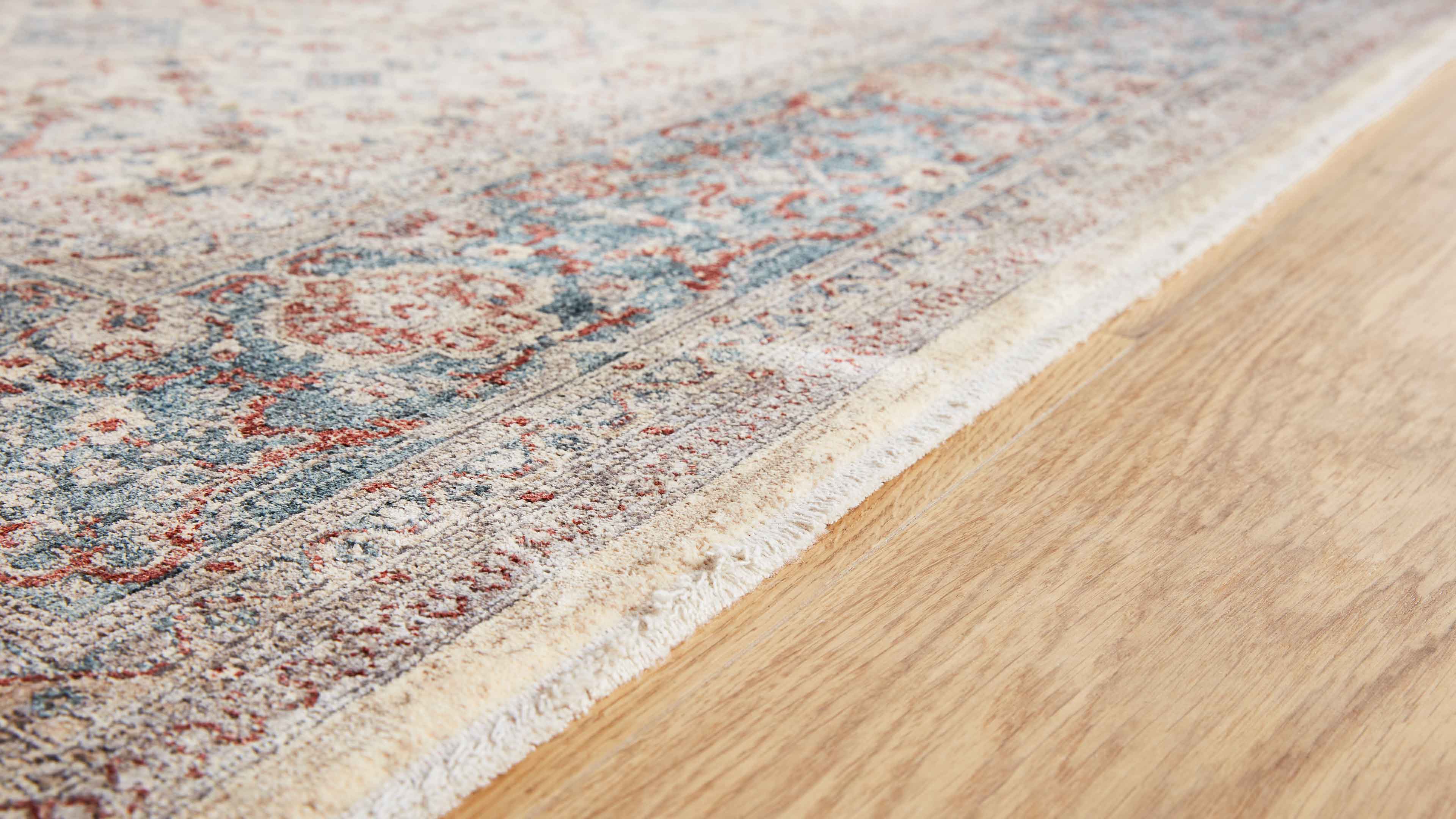 Teppich mit kurzem Flor, beigem Fransenrand und blau-roten Mustern liegt auf hellem Holzfußboden.