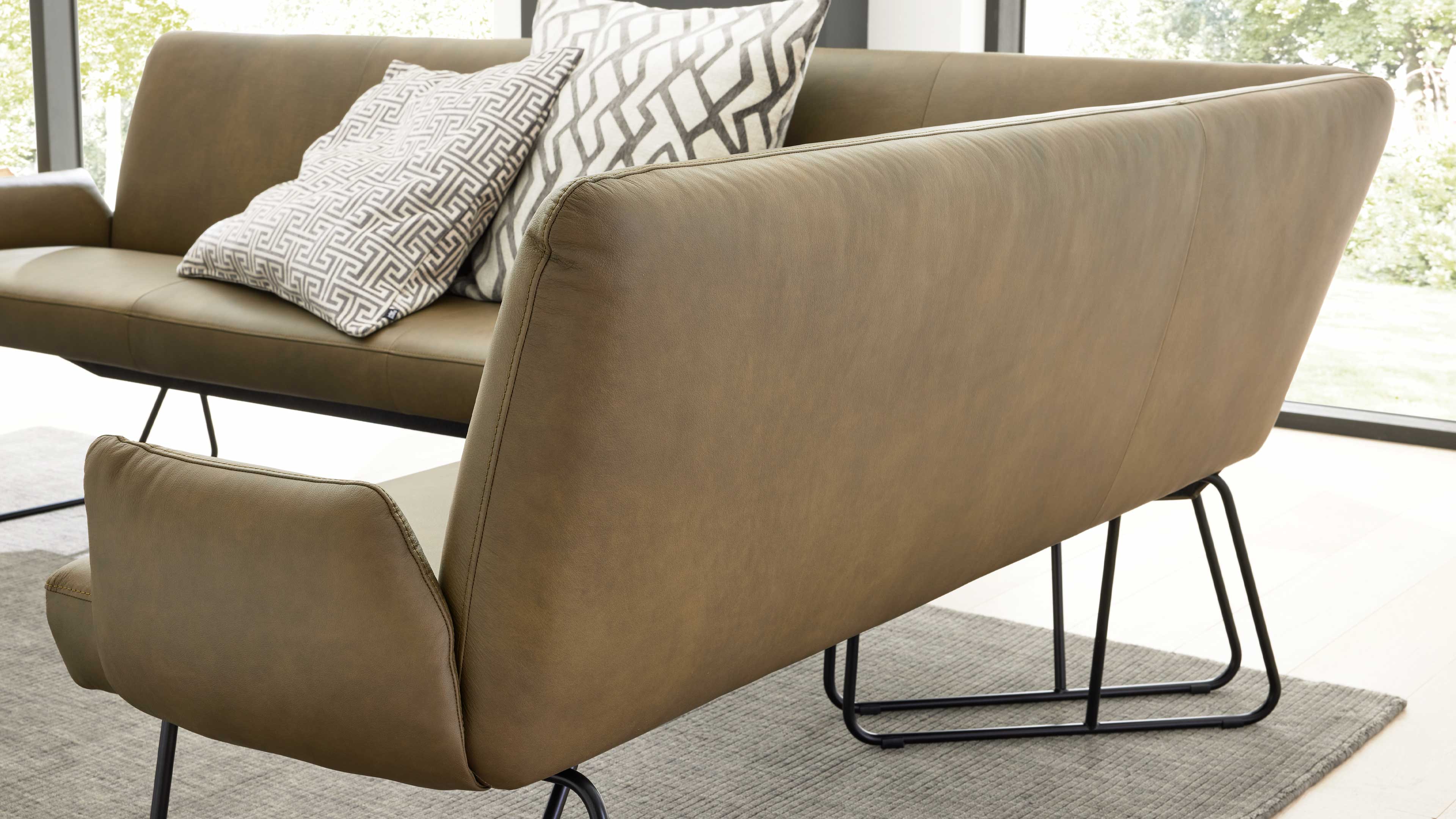 Hellbraunes Sofa mit abgerundeter Rückenlehne und schwarzen Metallkufen, dahinter weiteres Sofa mit zwei gemusterten Kissen vor Fenster.