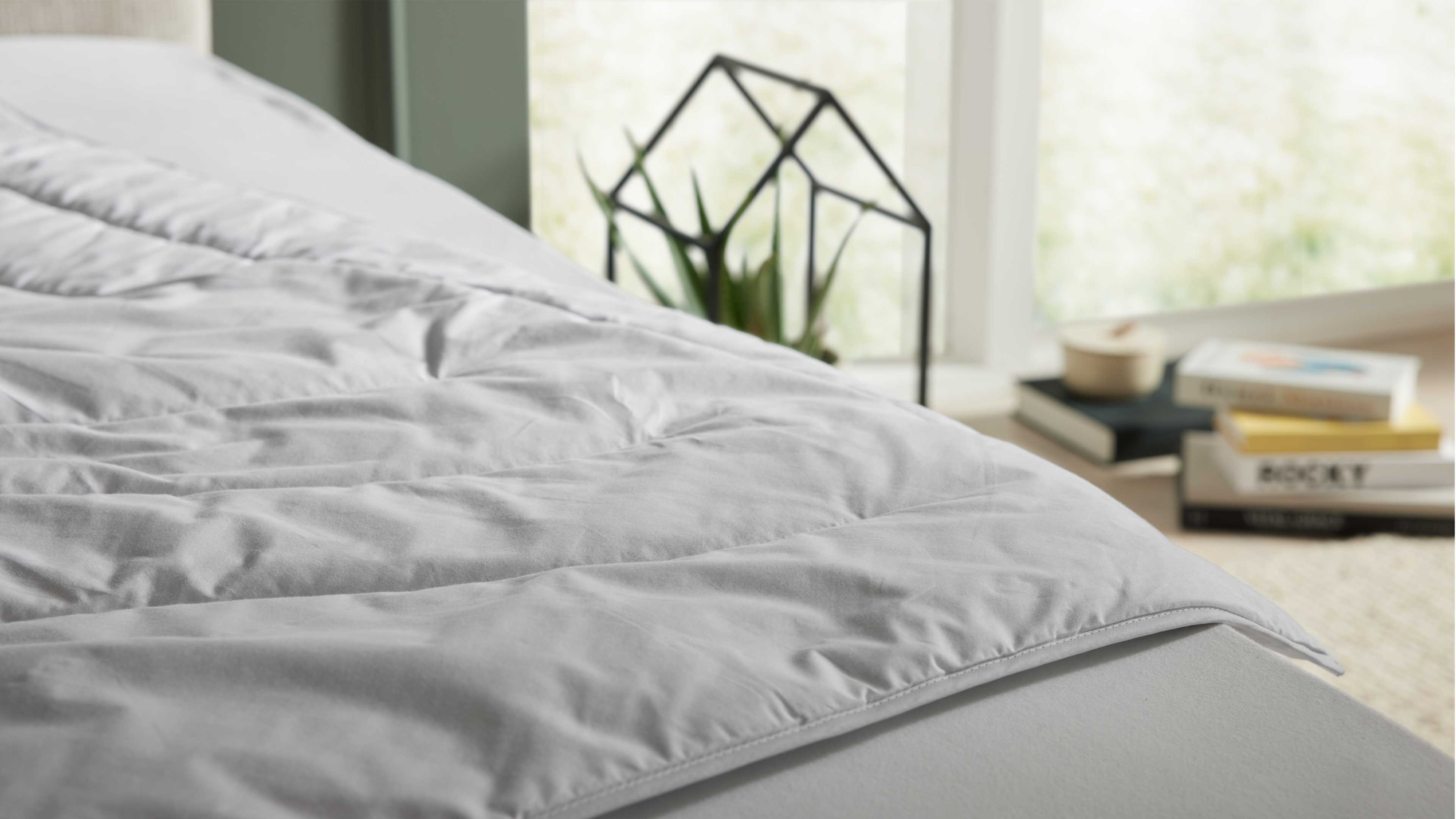 Grau-weiß gesteppte Bettdecke liegt über der Matratzenkante, im Hintergrund Fenster mit Pflanze im Metallgestell und gestapelten Büchern.