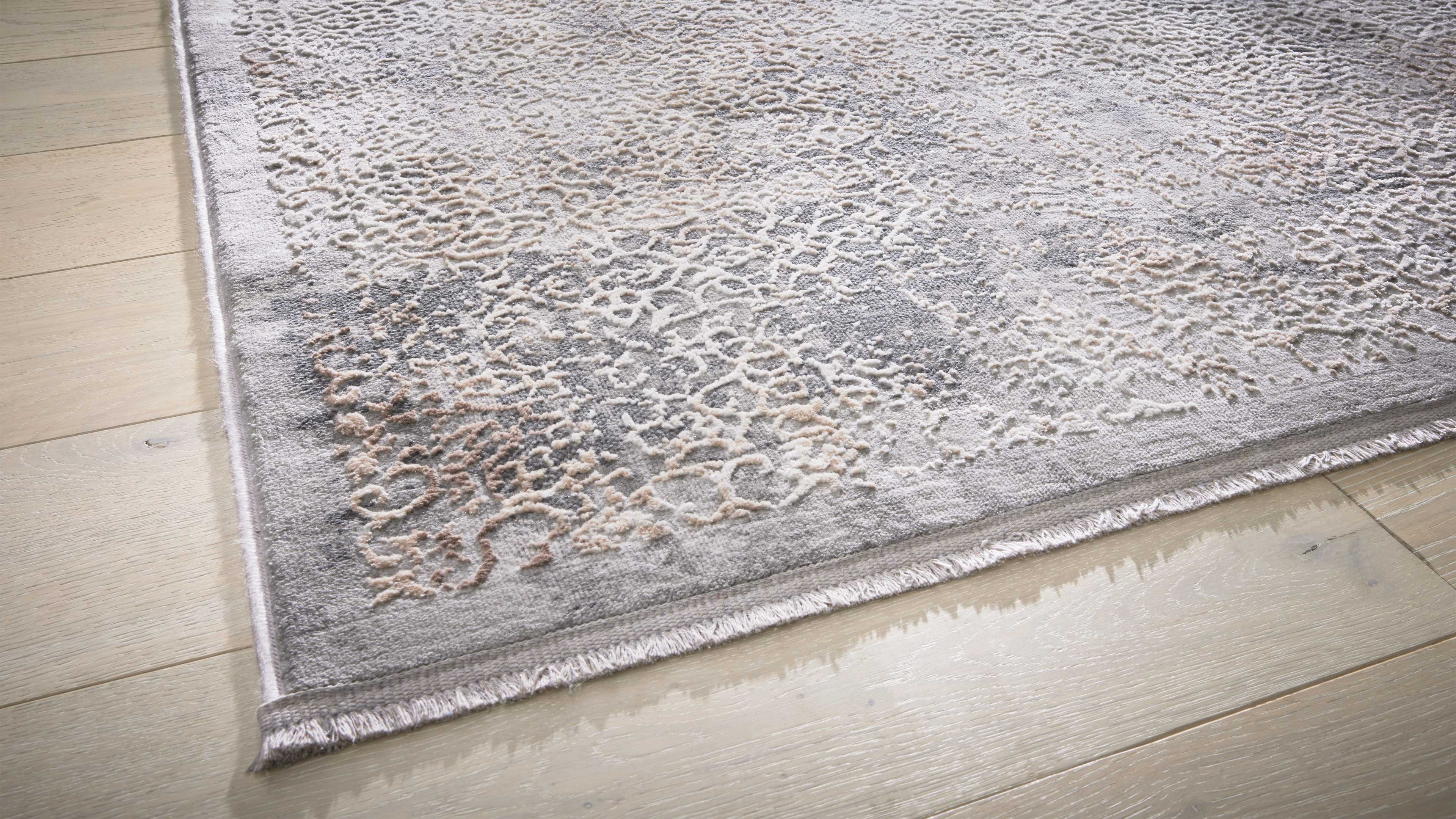 Eckansicht eines hellgrauen Teppichs mit reliefartigem beigen Muster und kurzem Fransenabschluss auf hellen Holzdielen.
