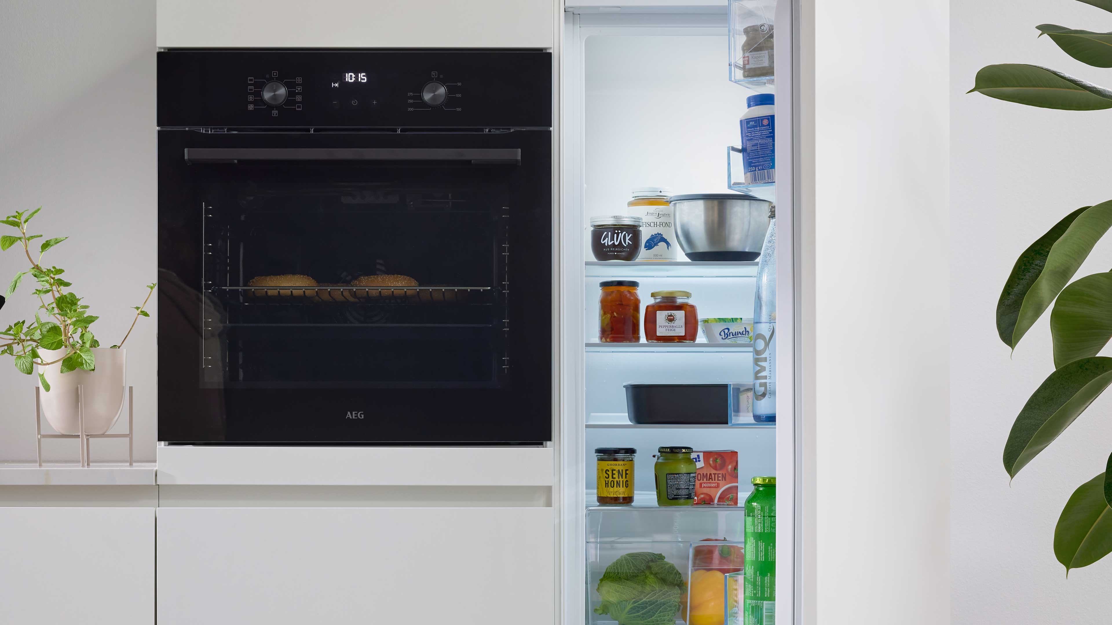 Eingebauter schwarzer Ofen mit zwei Backwaren auf dem Rost, daneben geöffneter Kühlschrank mit Gläsern, Schale und Flaschen.