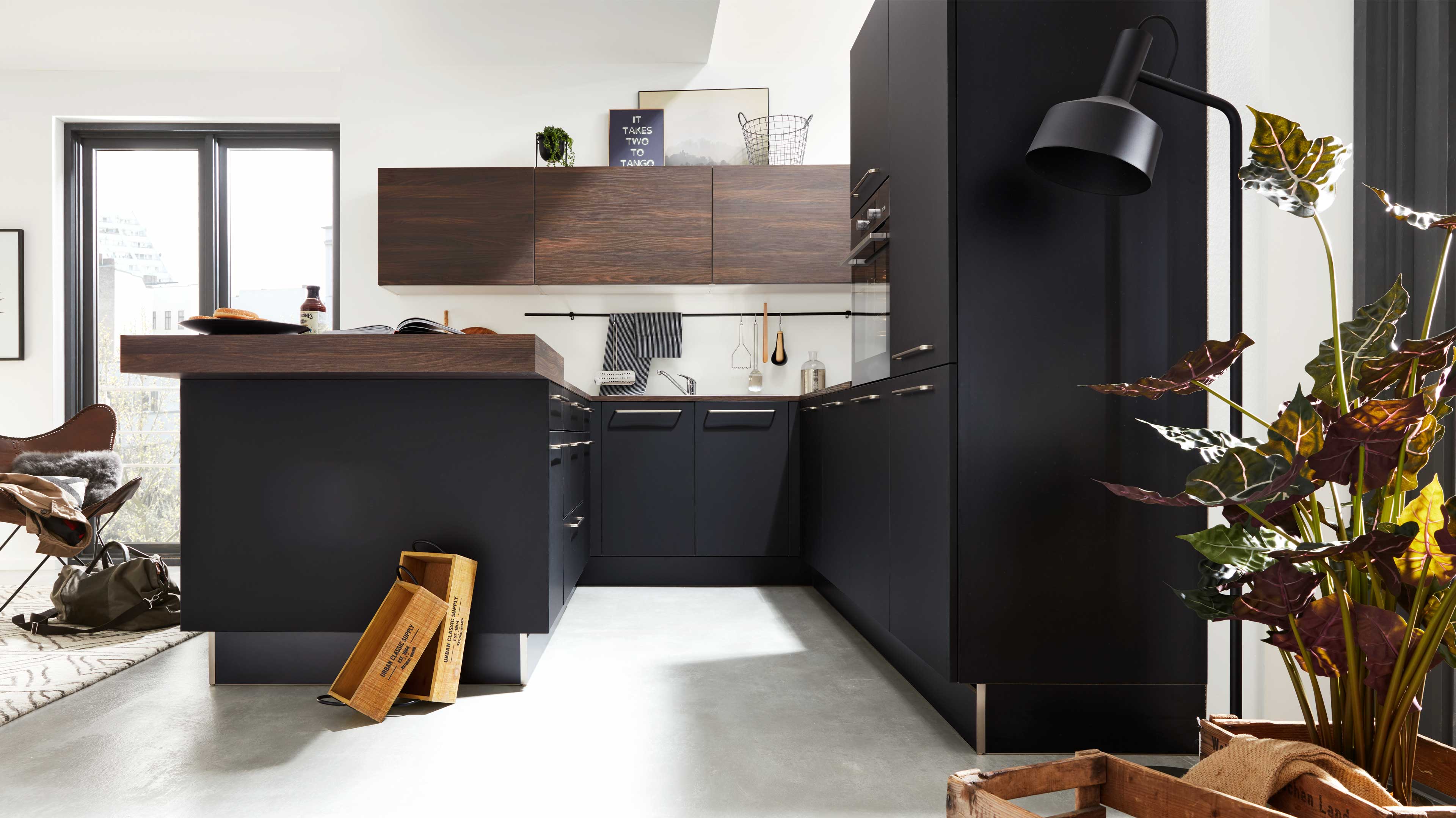Dunkle L-förmige Küchenmöbel mit massiver Holzarbeitsplatte, Hängeschränken und Insel, rechts große Topfpflanze, links Fenster.