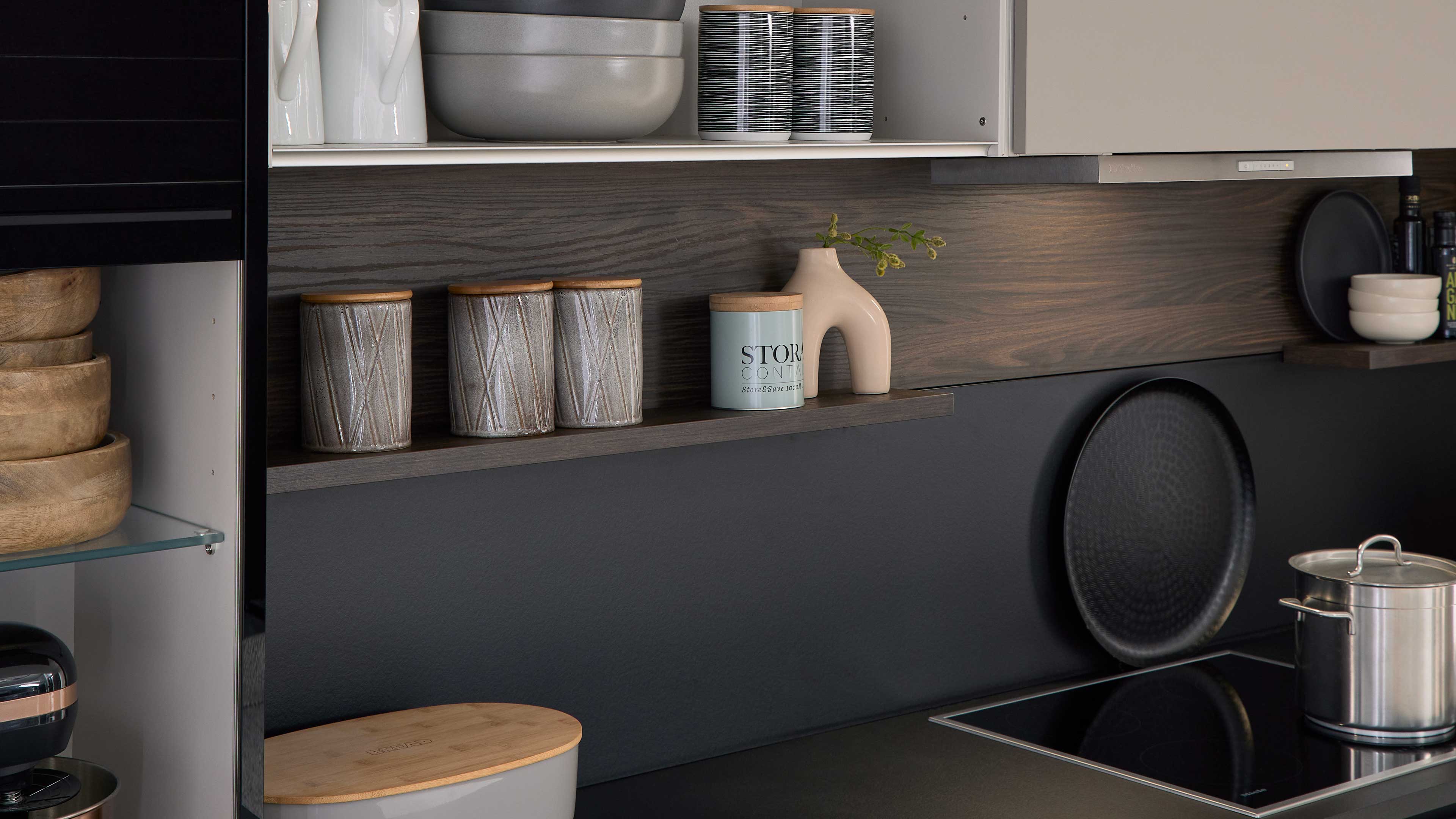 Wandregal über Kochfeld mit dunkler Holzrückwand, drei graue Keramikbehälter mit Holzdeckeln, kleine Dose, beige Vase und Kochtopf.