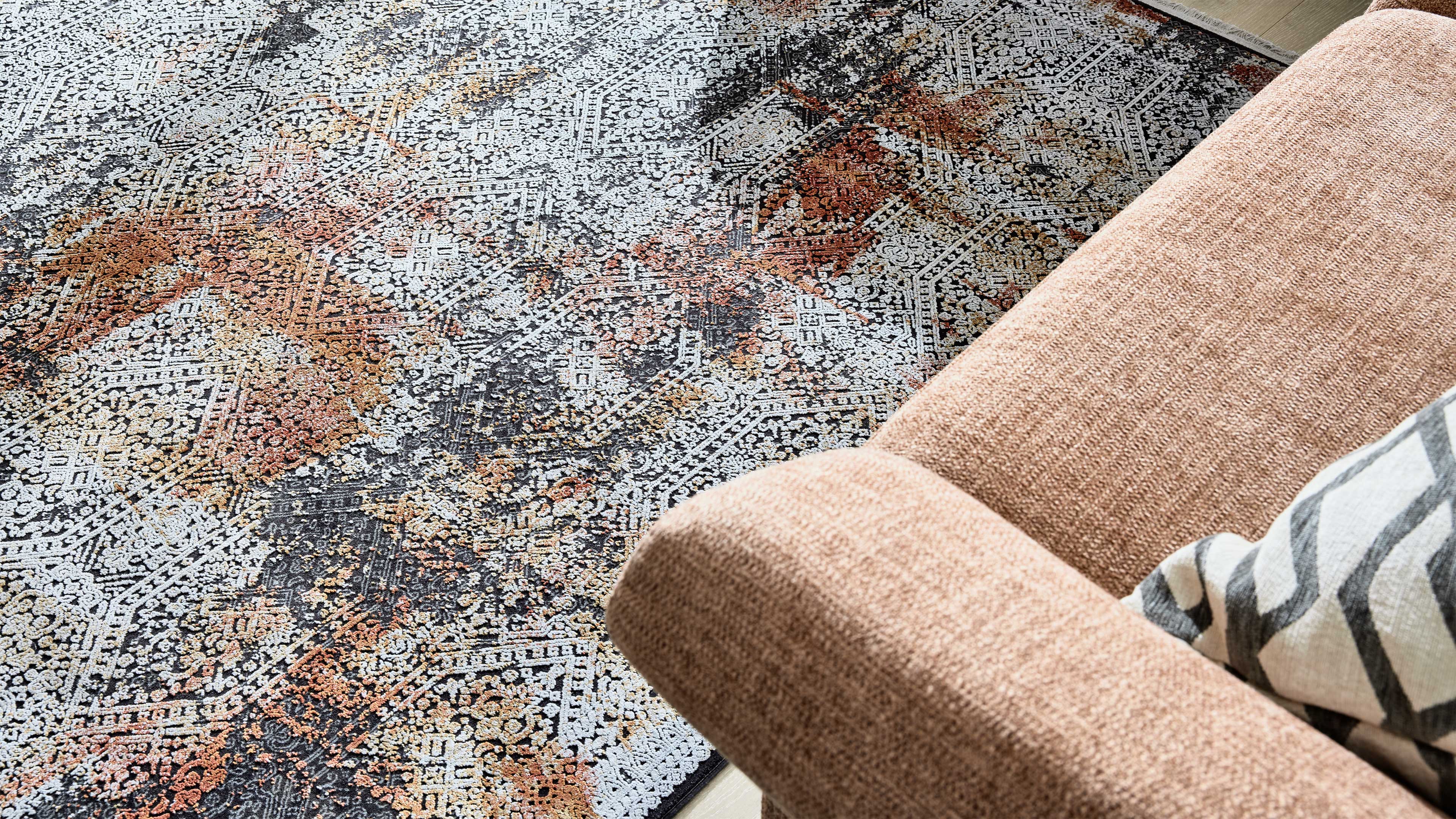 Großflächiger Teppich mit geometrisch‑floralem Muster in Grau, Weiß und Rosttönen, daneben hellrosa Sofaarm mit gestreiftem Kissen.