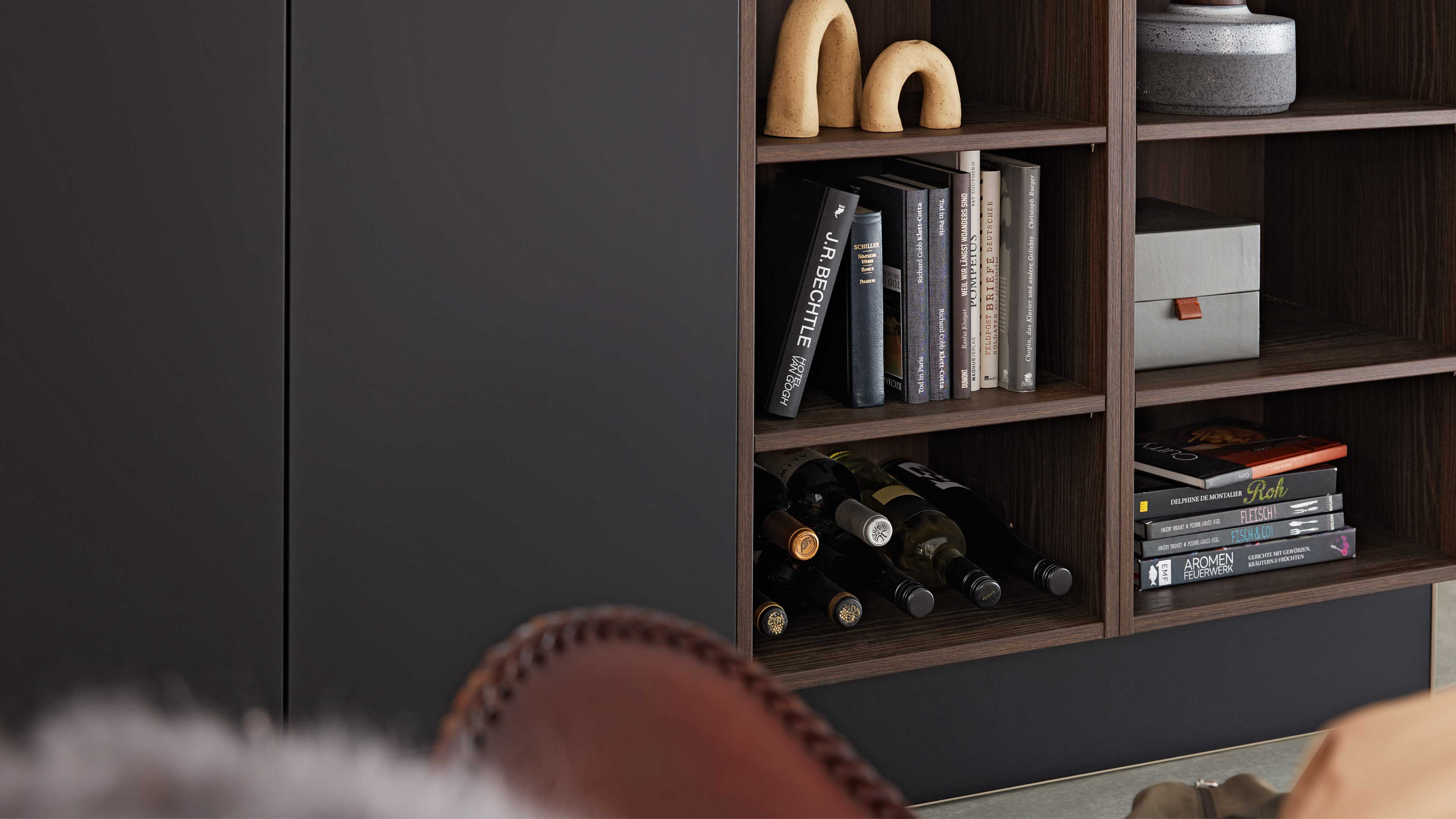 Offenes dunkles Holzregal mit Büchern, liegenden Weinflaschen, zwei bogenförmigen Keramikfiguren oben und grauer Box rechts.