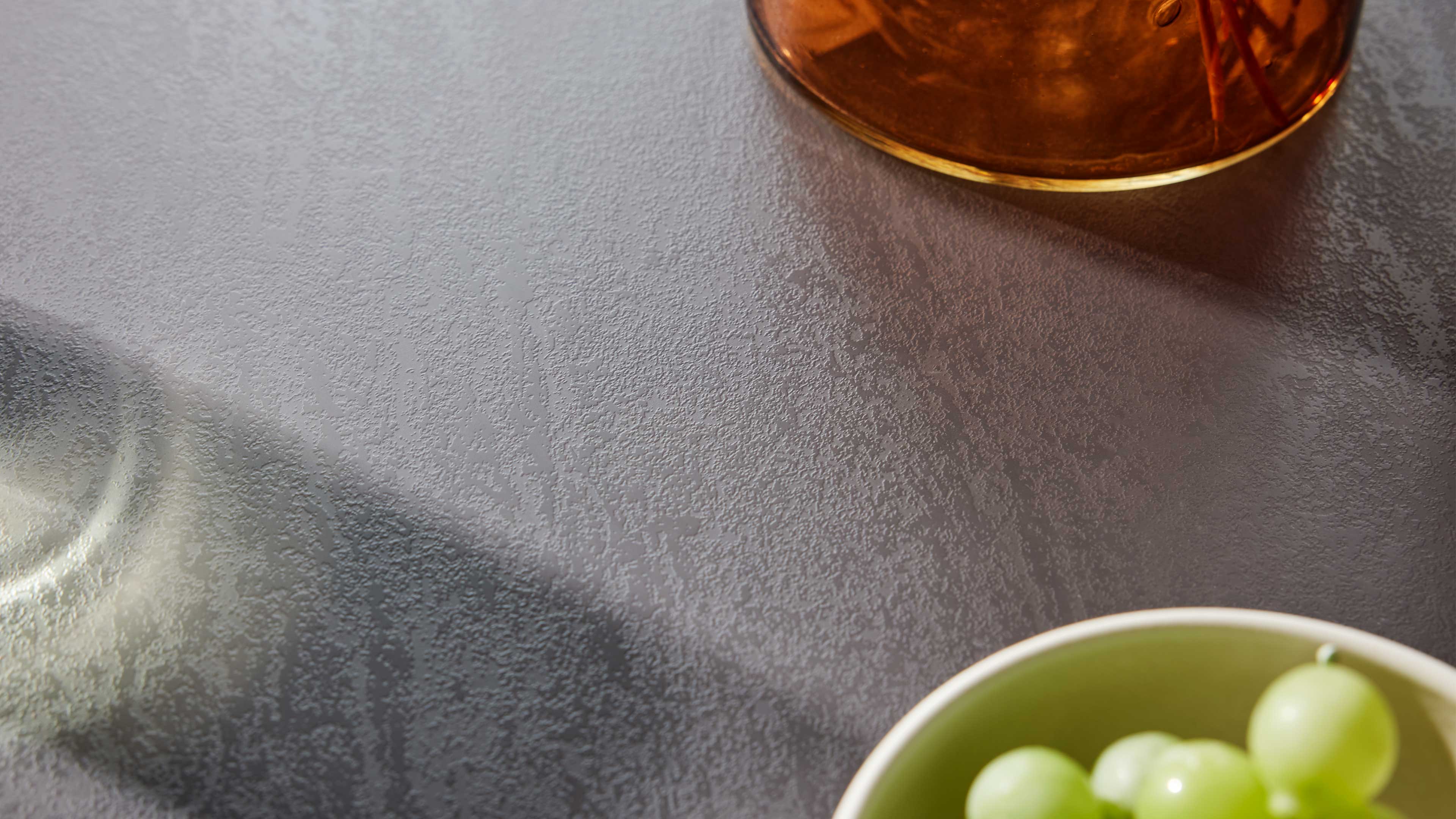 Grau strukturierte Tischfläche mit Schatten, rechts oben Teil eines bernsteinfarbenen Glasgefäßes und unten Schüsselrand mit grünen Trauben.