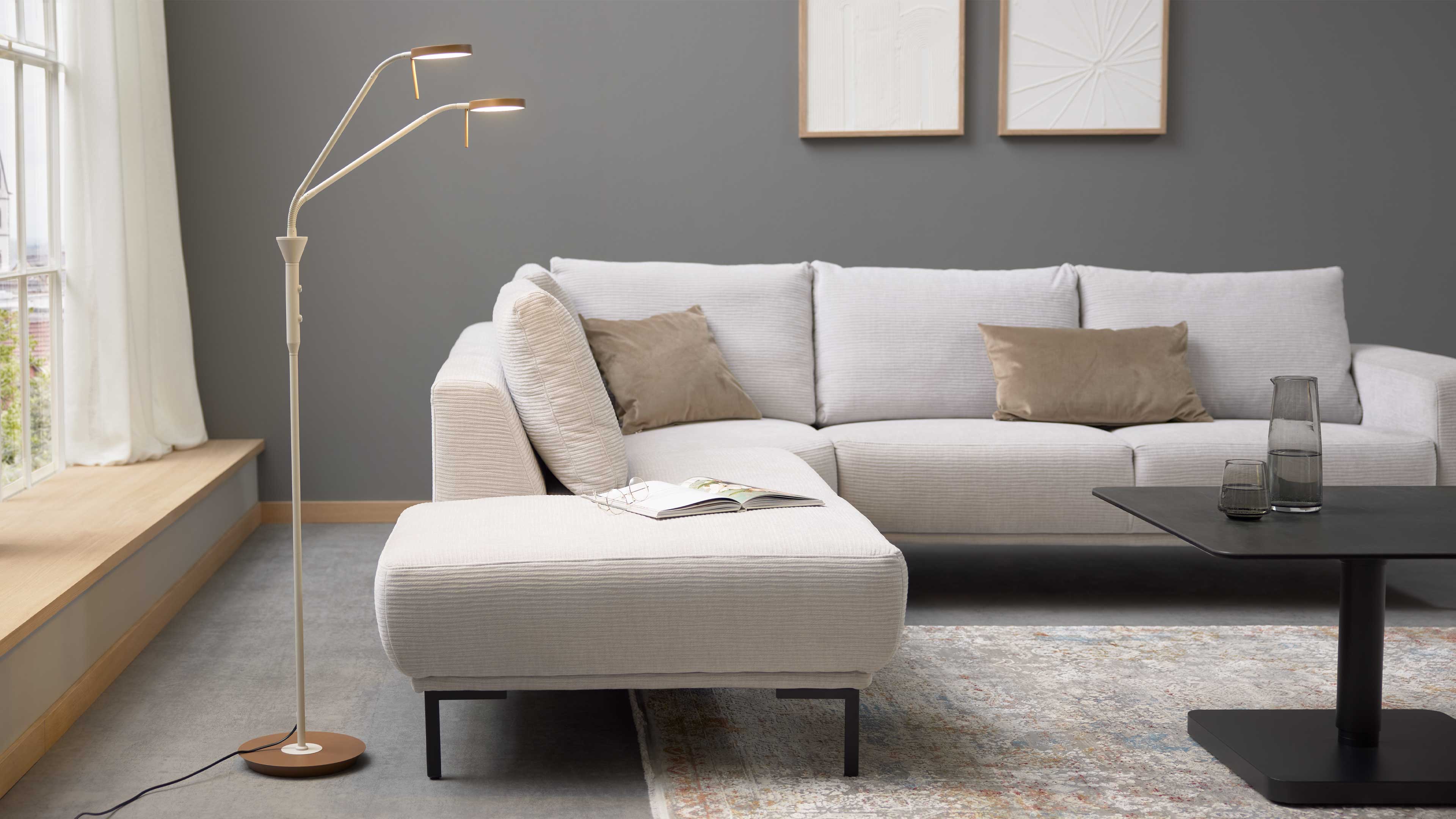 Beiges Ecksofa mit Chaiselongue, zwei braunen Kissen und offenem Magazin, links Stehleuchte, rechts schwarzer Couchtisch mit Glasvase.