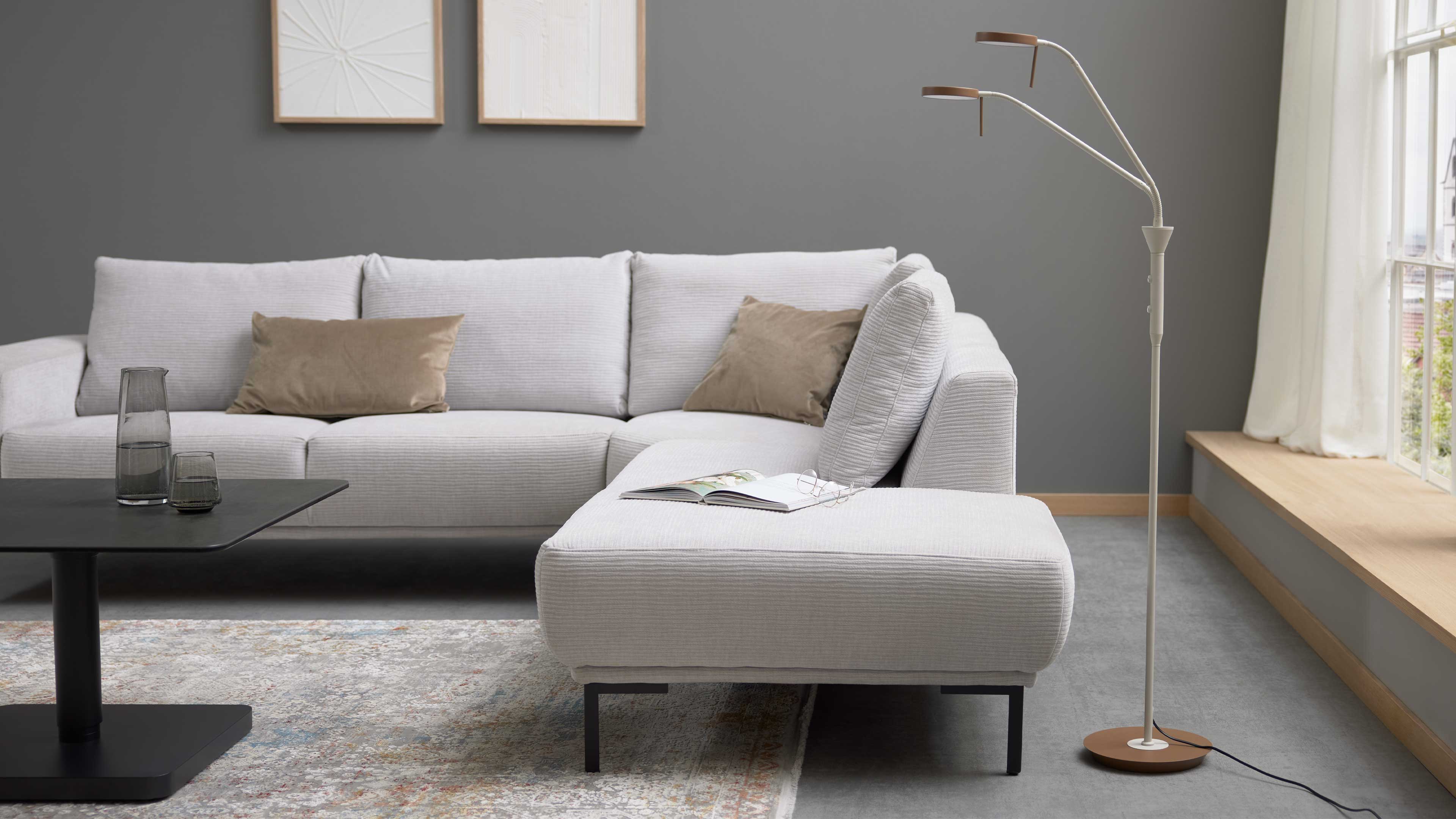 Helles, hellgraues Ecksofa mit Ottomane, zwei beige Kissen, schwarzem Couchtisch und dreiflammiger Stehleuchte.