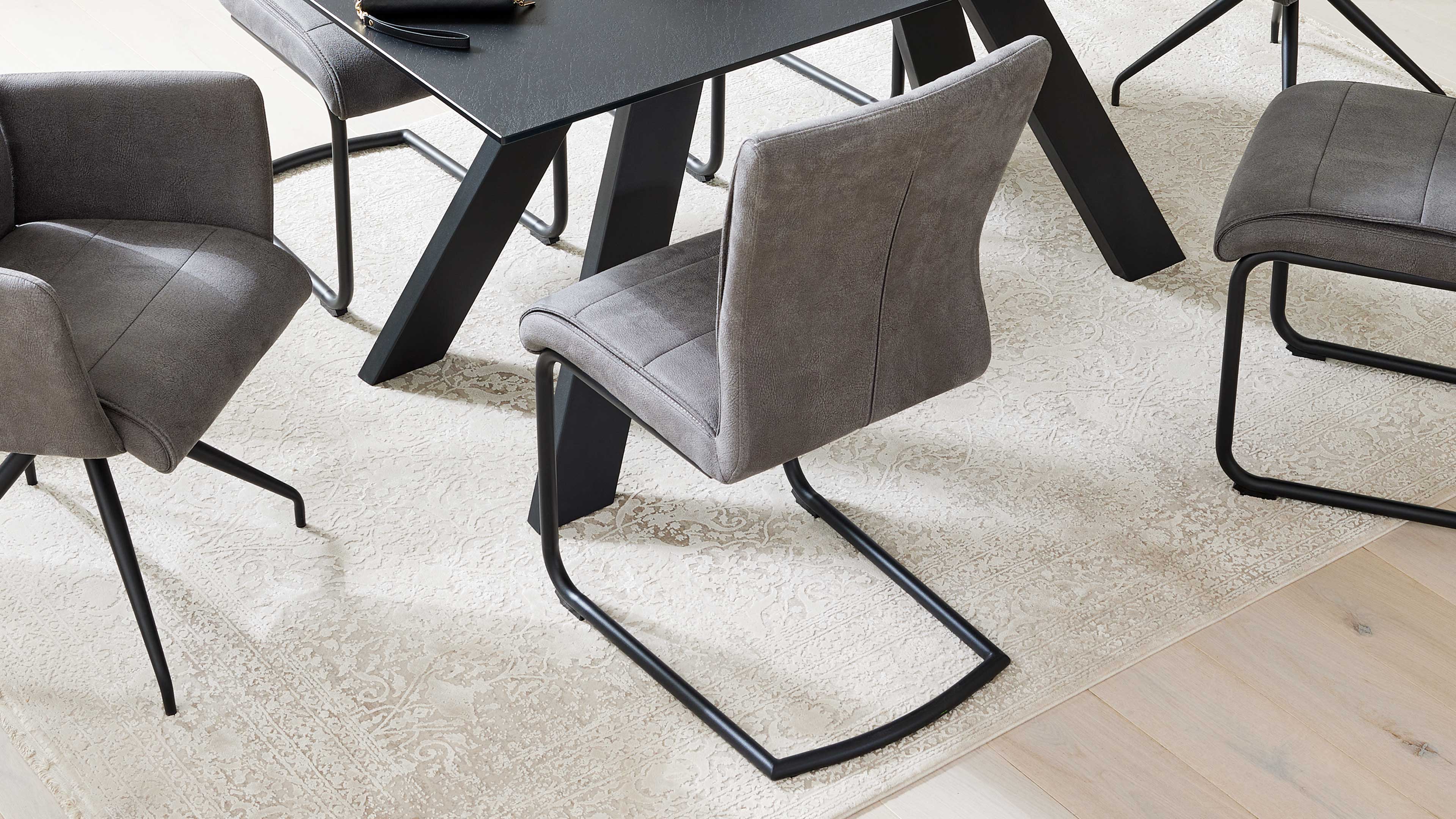 Grau gepolsterter Freischwinger-Stuhl mit schwarzem Metallgestell vor schwarzem Tisch und weiteren Stühlen auf hellgemusteretem Teppich.