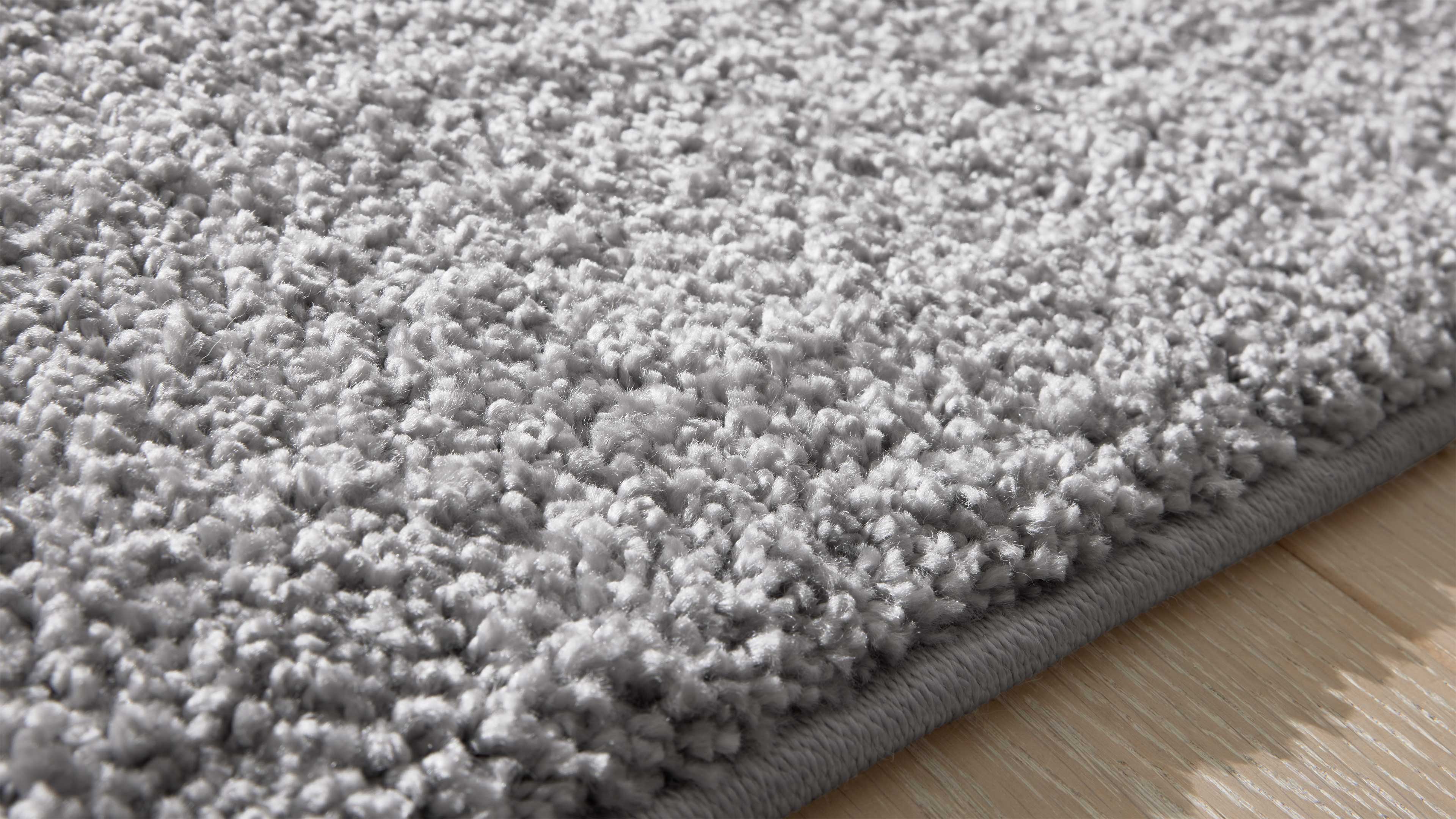 Nahaufnahme eines hellgrauen, flauschigen Hochflorteppichs mit eingefasster Kante auf hellem Holzfußboden.