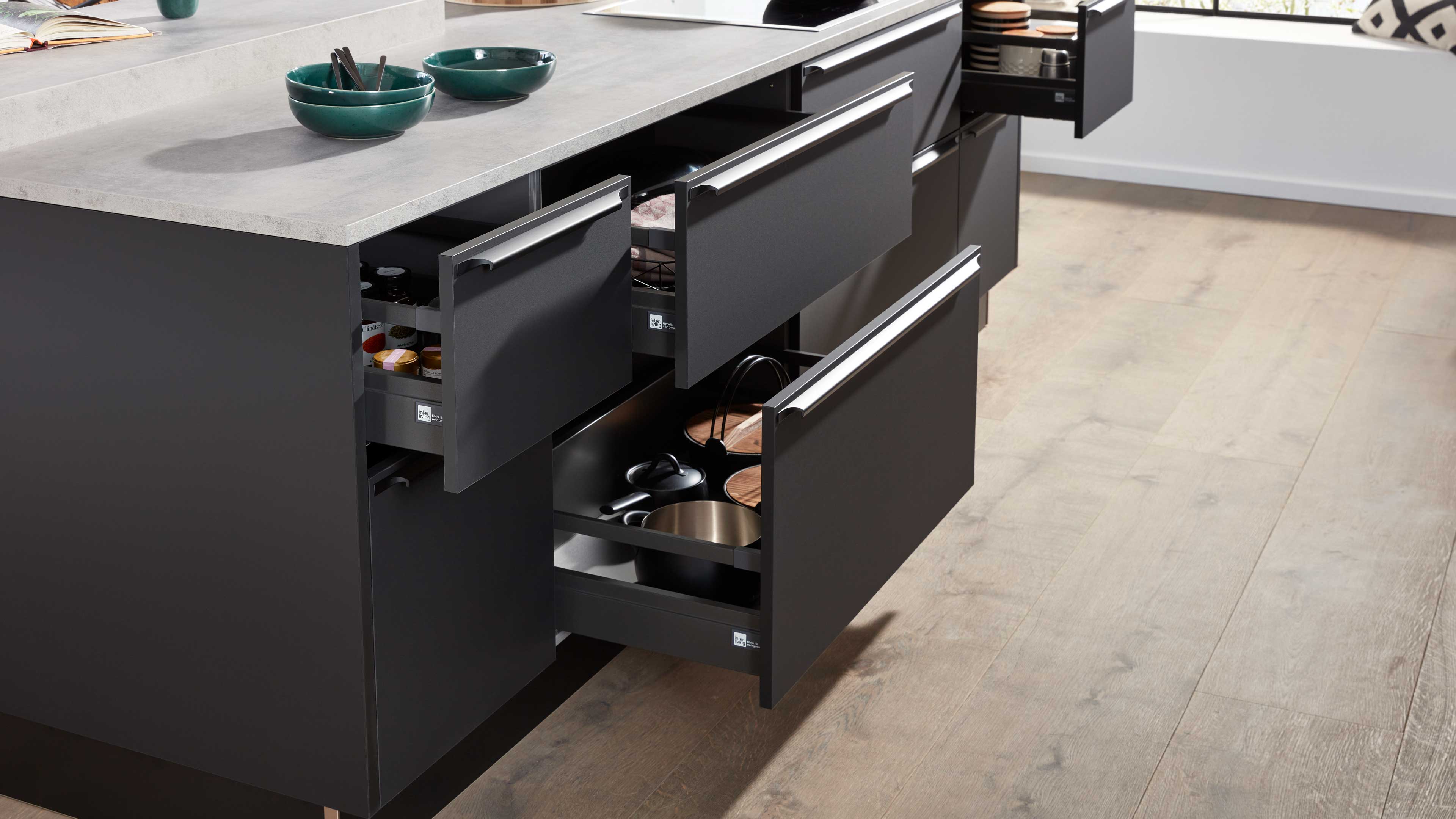 Offene dunkelgraue Küchenschubladen unter hellem Steinarbeitsplatte, gefüllt mit Töpfen, Deckeln, Gewürzgläsern und grünen Schalen oben.