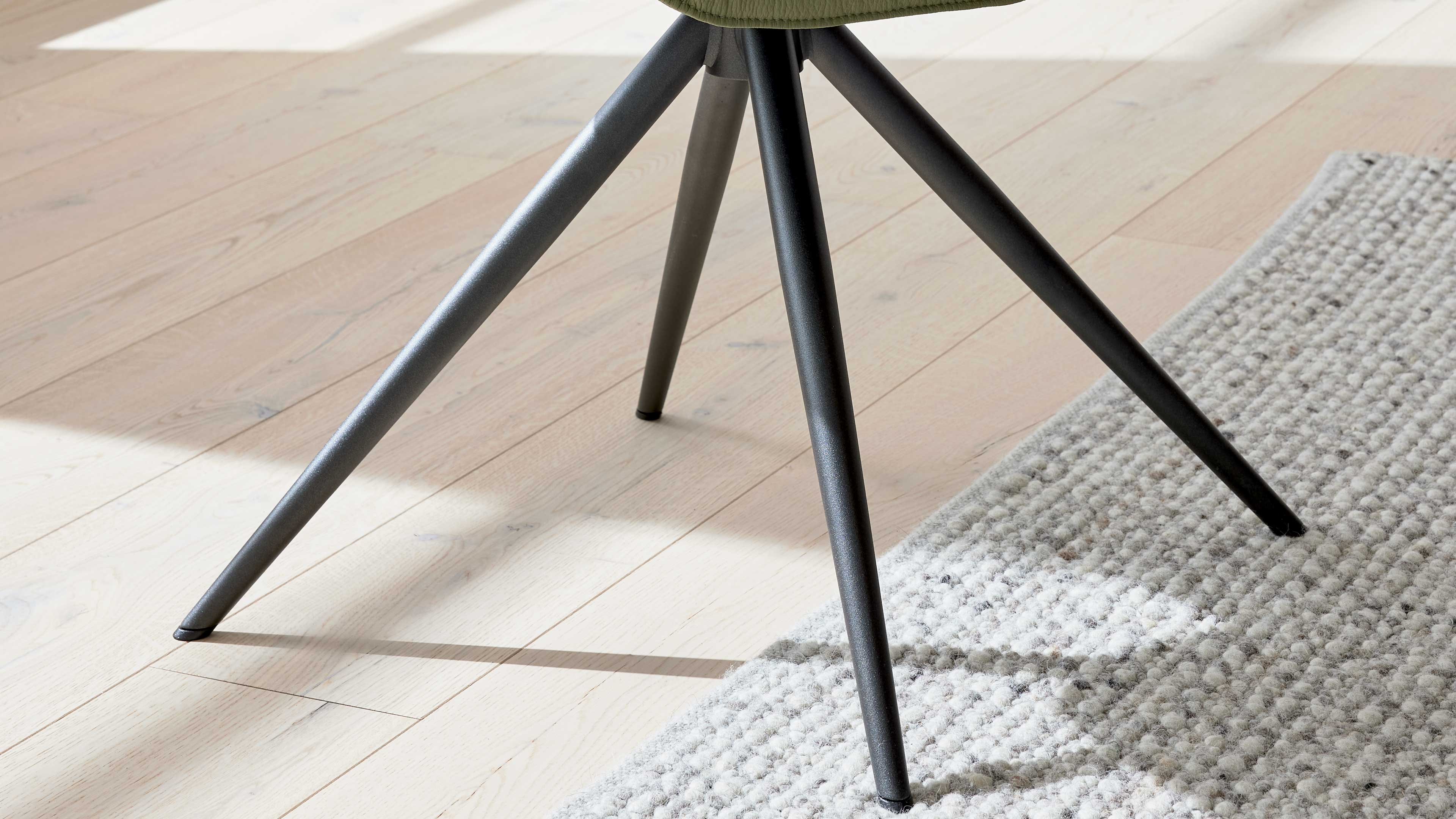 Drei schräg gestellte, dunkle Metallbeine eines Stuhls treffen in der Mitte, auf hellem Parkettboden neben hellem grauen Teppich.