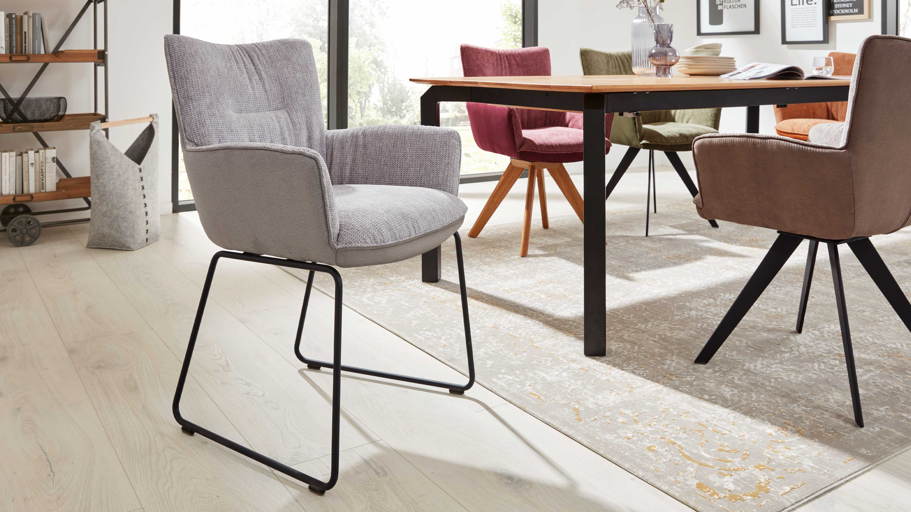 Grauer gepolsterter Armlehnstuhl mit schwarzem Metallgestell, davor Holztisch auf Teppich und bunte Essstühle.