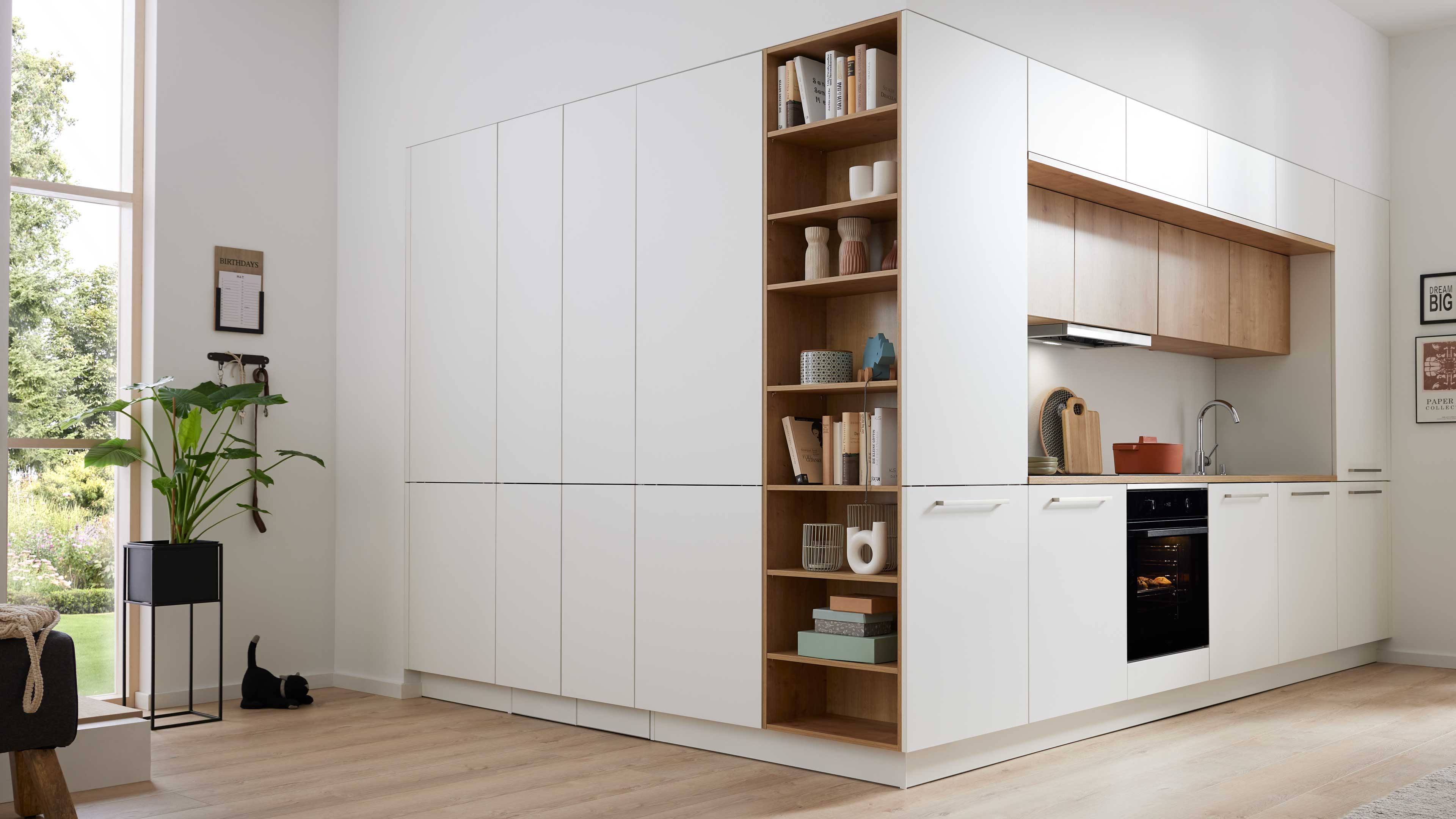 Weiße Küchenzeile mit offenem Holzregal, eingebautem Backofen und Spüle, rechts Oberschränke; links großes Fenster mit grüner Topfpflanze.