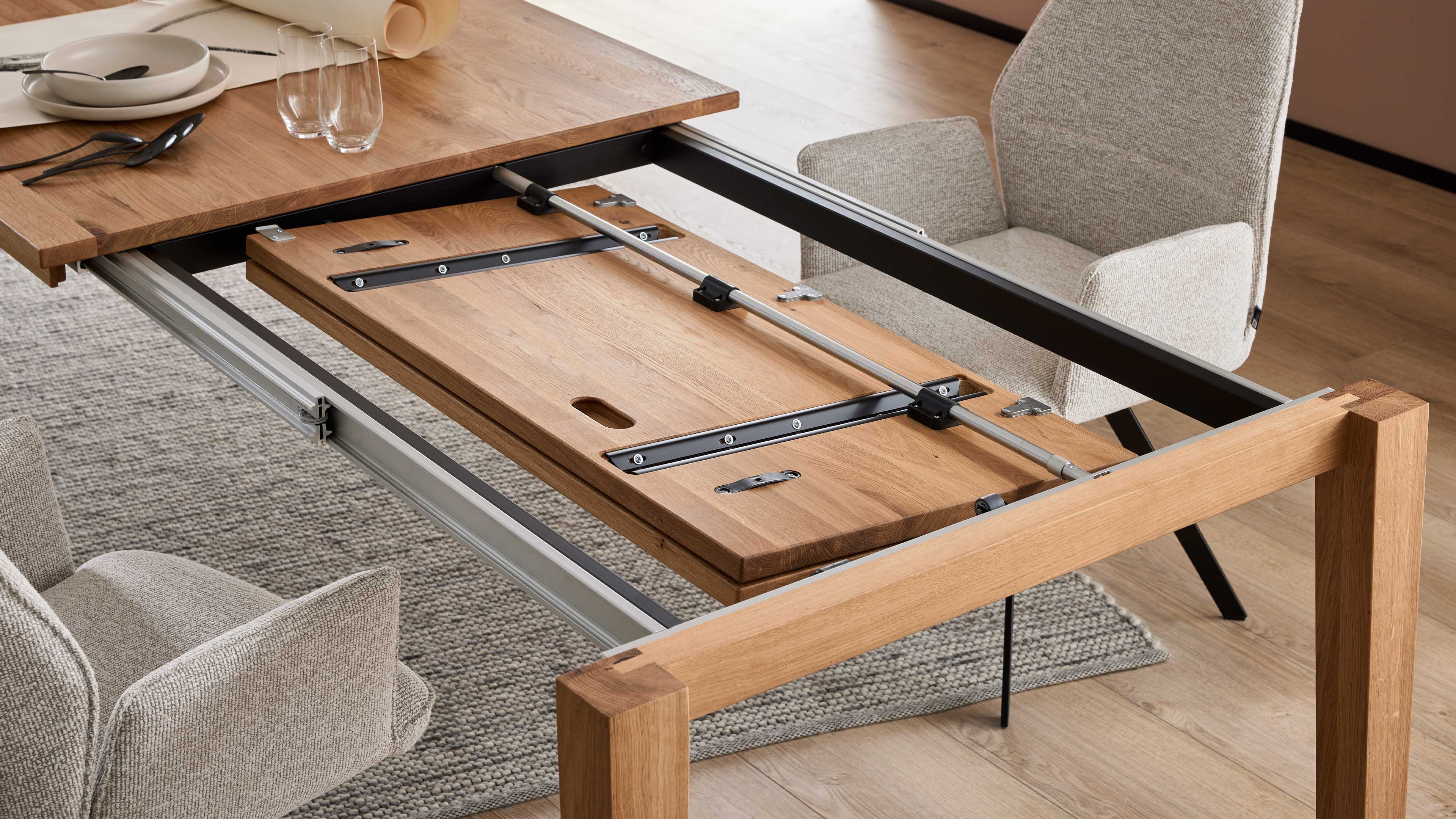 Ausgezogener Esstisch mit geöffneter Holz-Einlegeplatte, sichtbaren Metallführungen und zwei hellen, gepolsterten Stühlen.