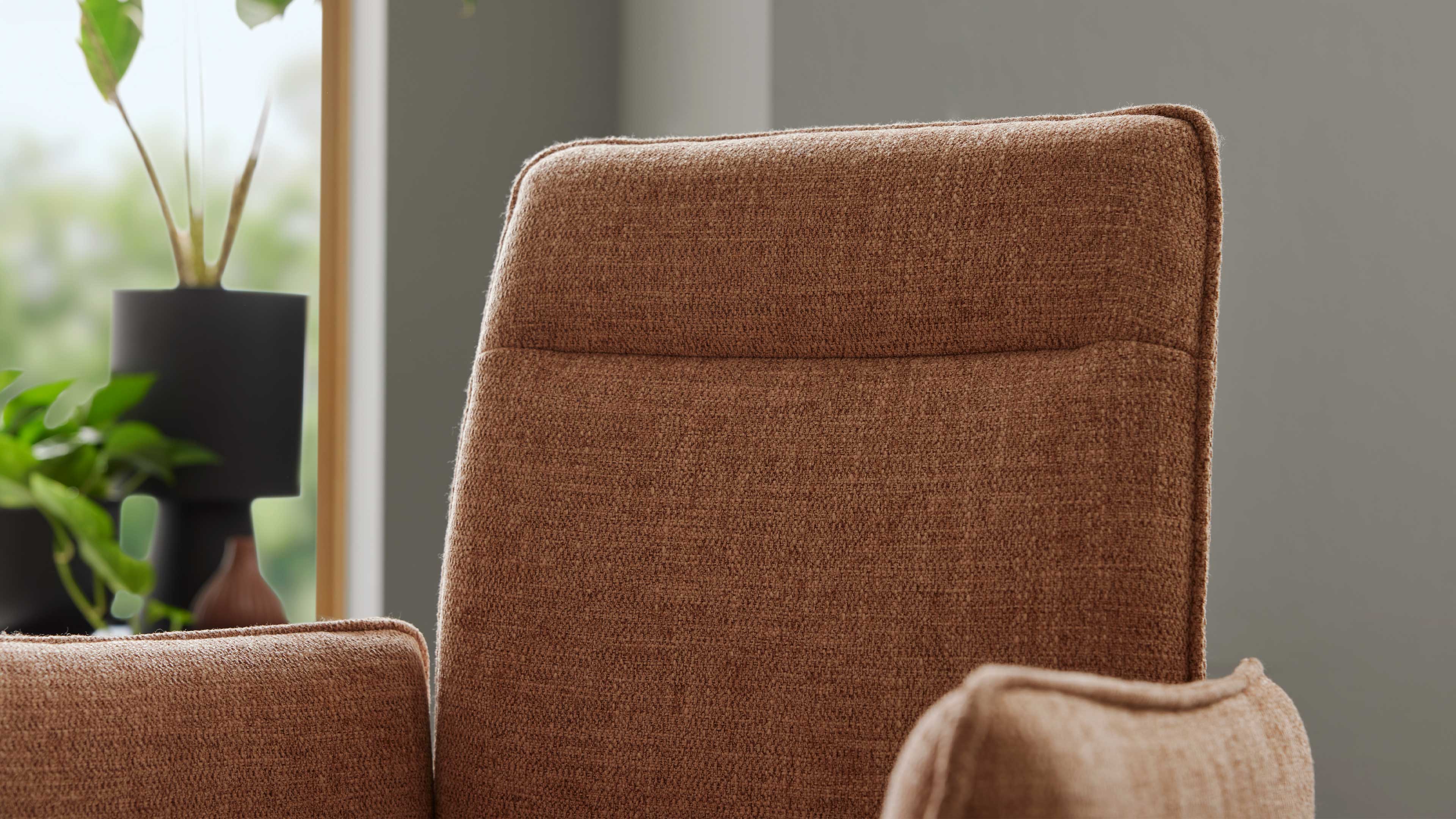 Braunes, grobgewebtes Polster eines Sessels mit hoher Rückenlehne und sichtbarem Armkissen, im Hintergrund Pflanzen und graue Wand.