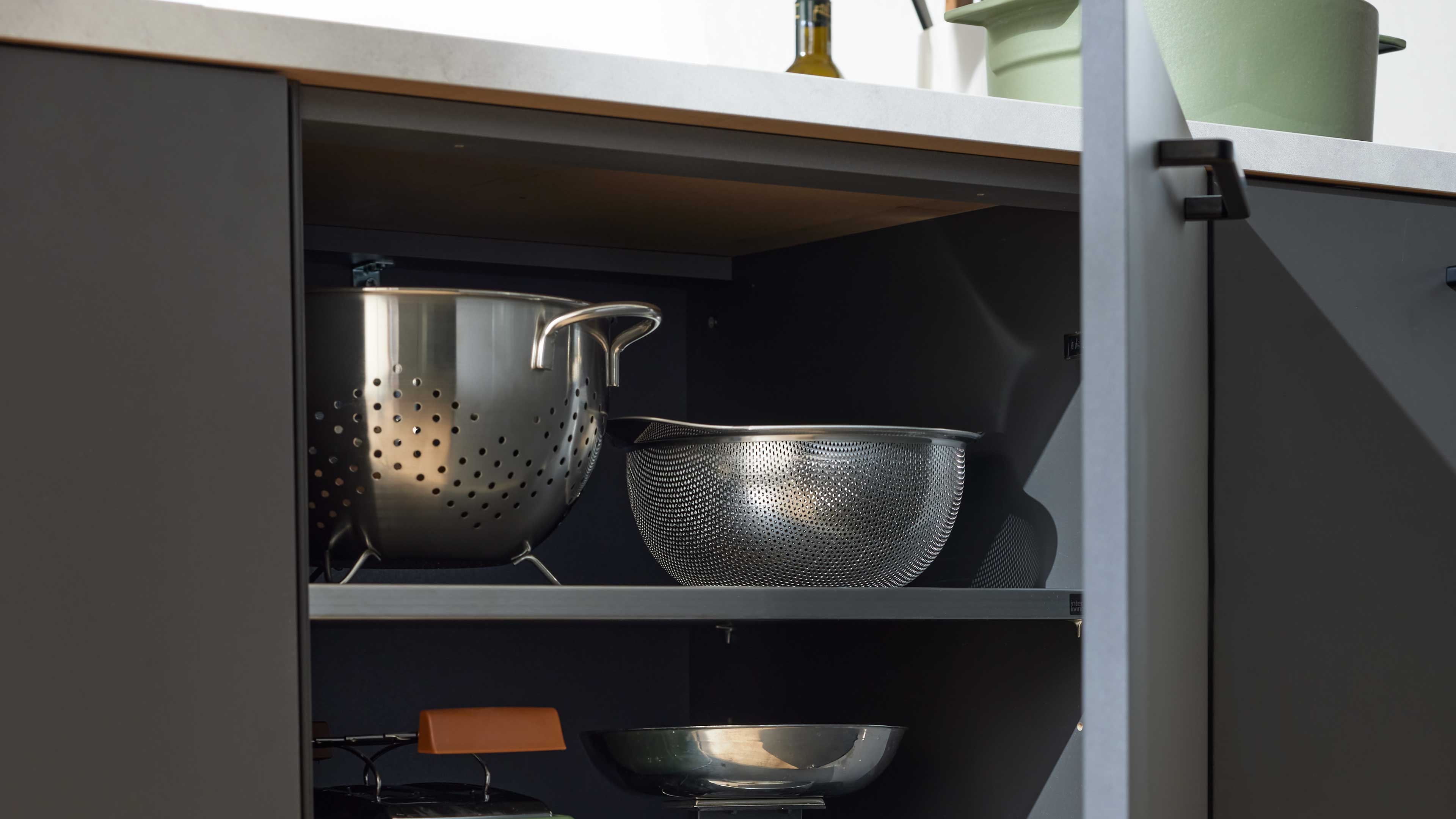 Offener dunkelgrauer Küchenschrank mit zwei Edelstahlsieben auf einem Regalbrett und weiteren Küchenutensilien.