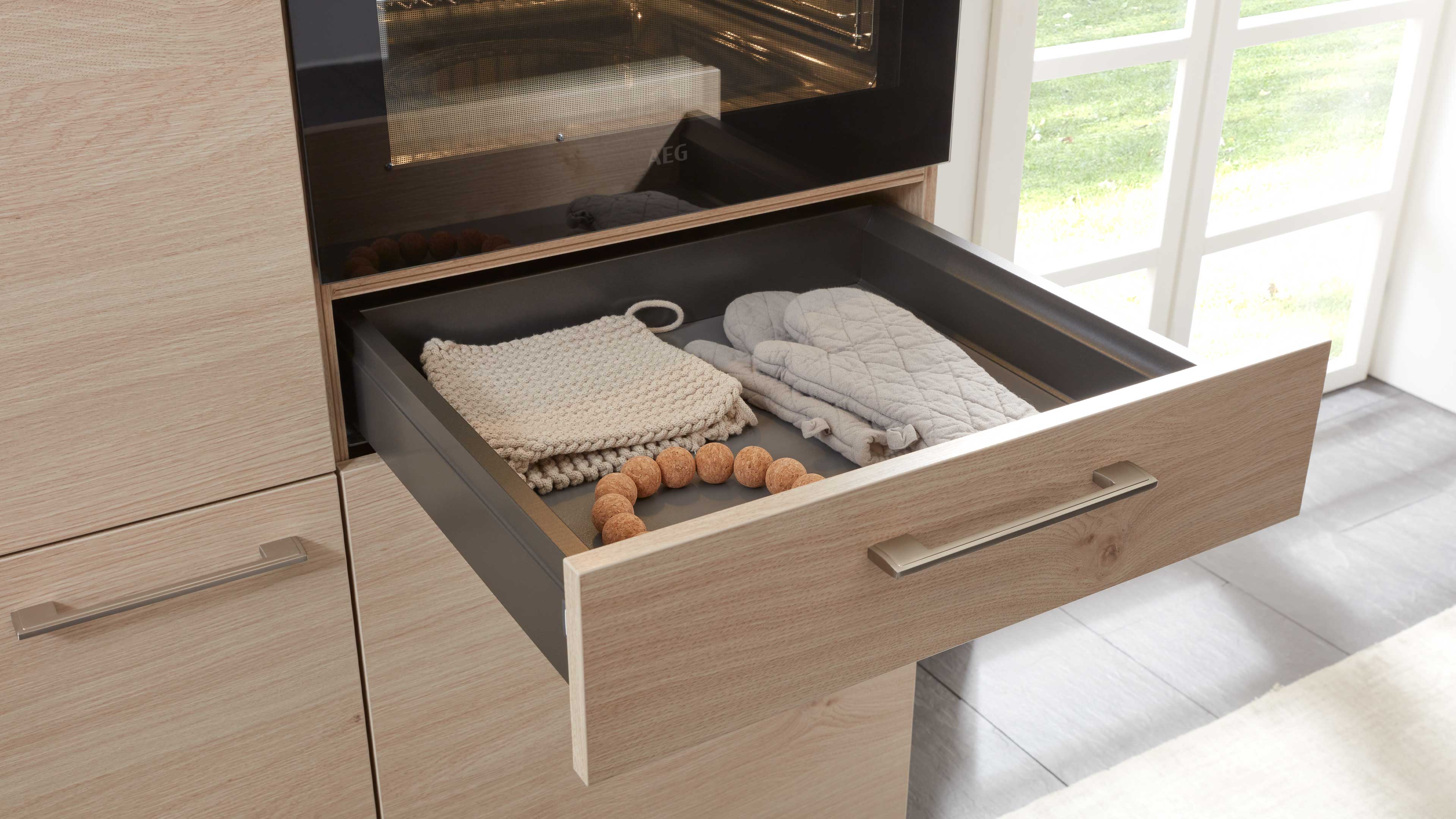 Ausgezogene Schublade mit hellholzfront und metallgriff, dunkelgrauem Innenraum, darin Topflappen, Ofenhandschuhe und Korkperlen.