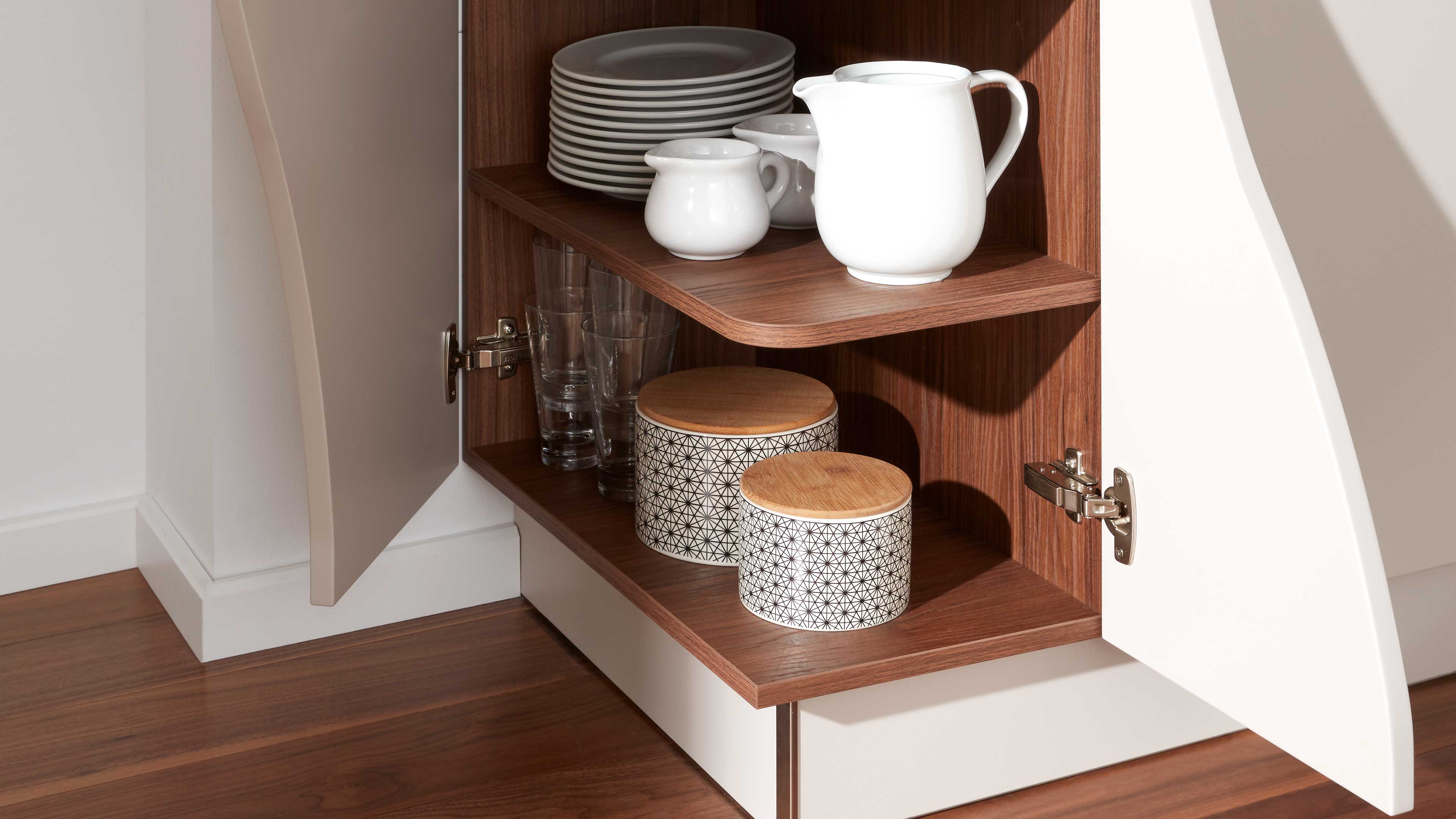 Offener Unterschrank mit weißen geschwungenen Türen und walnussfarbenen Böden, mit Keramikkannen, Tellern, Gläsern und zwei Dosen.