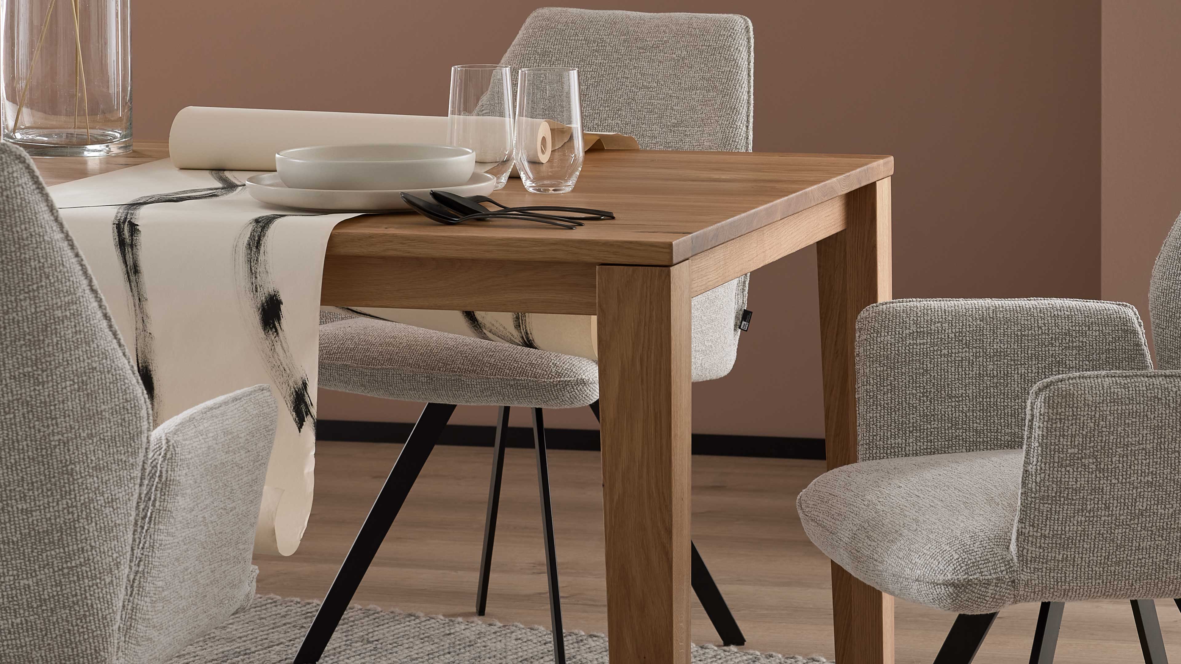 Rechteckiger Eichenholztisch mit hellem Läufer mit schwarzen Pinselstrichen, Tellern, zwei Gläsern und Besteck, umgeben von grauen Stühlen.