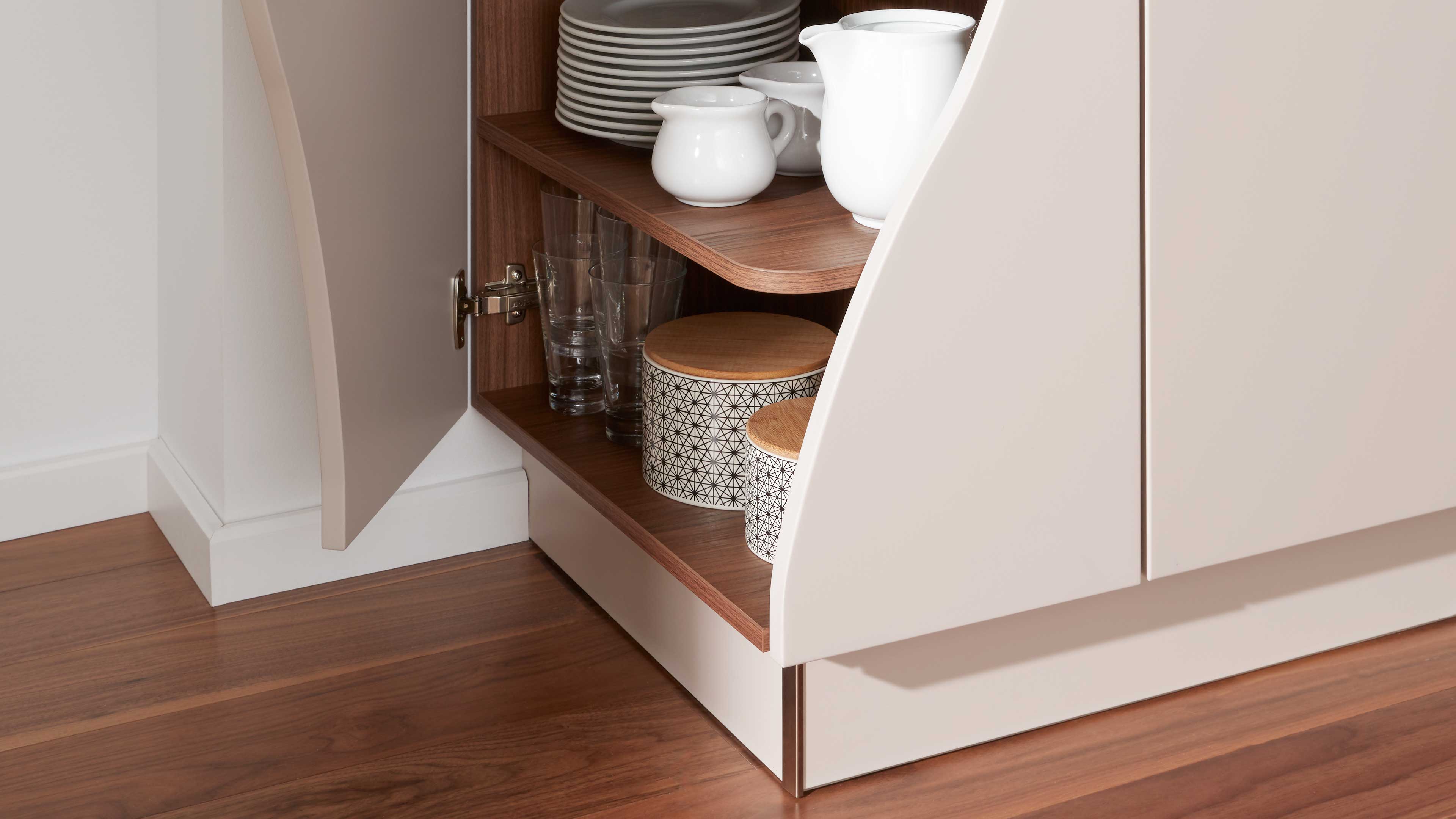 Geöffneter Küchenunterschrank mit hellbeigen geschwungenen Türen, Holzregalböden, Keramik‑ und Glasgeschirr sowie gemusterten Vorratsdosen.