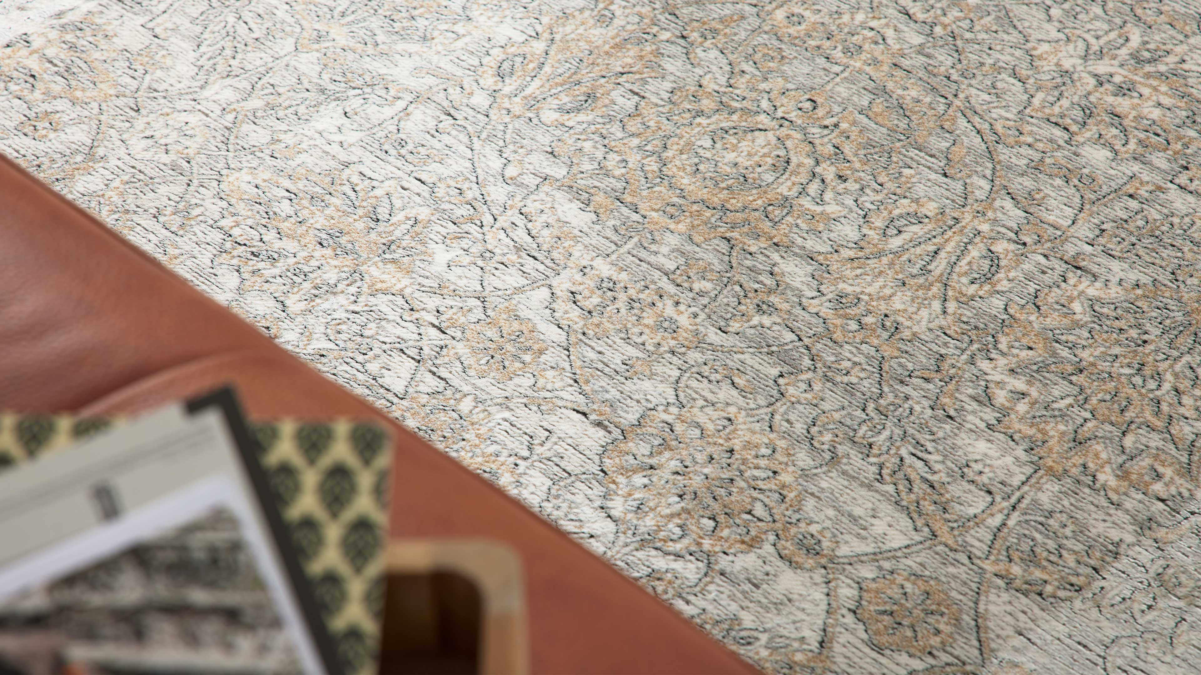Großflächiger hellbeiger Teppich mit floralen grauen und sandfarbenen Ornamenten, vorne braune Lederkante und unscharfe Zeitschriften.