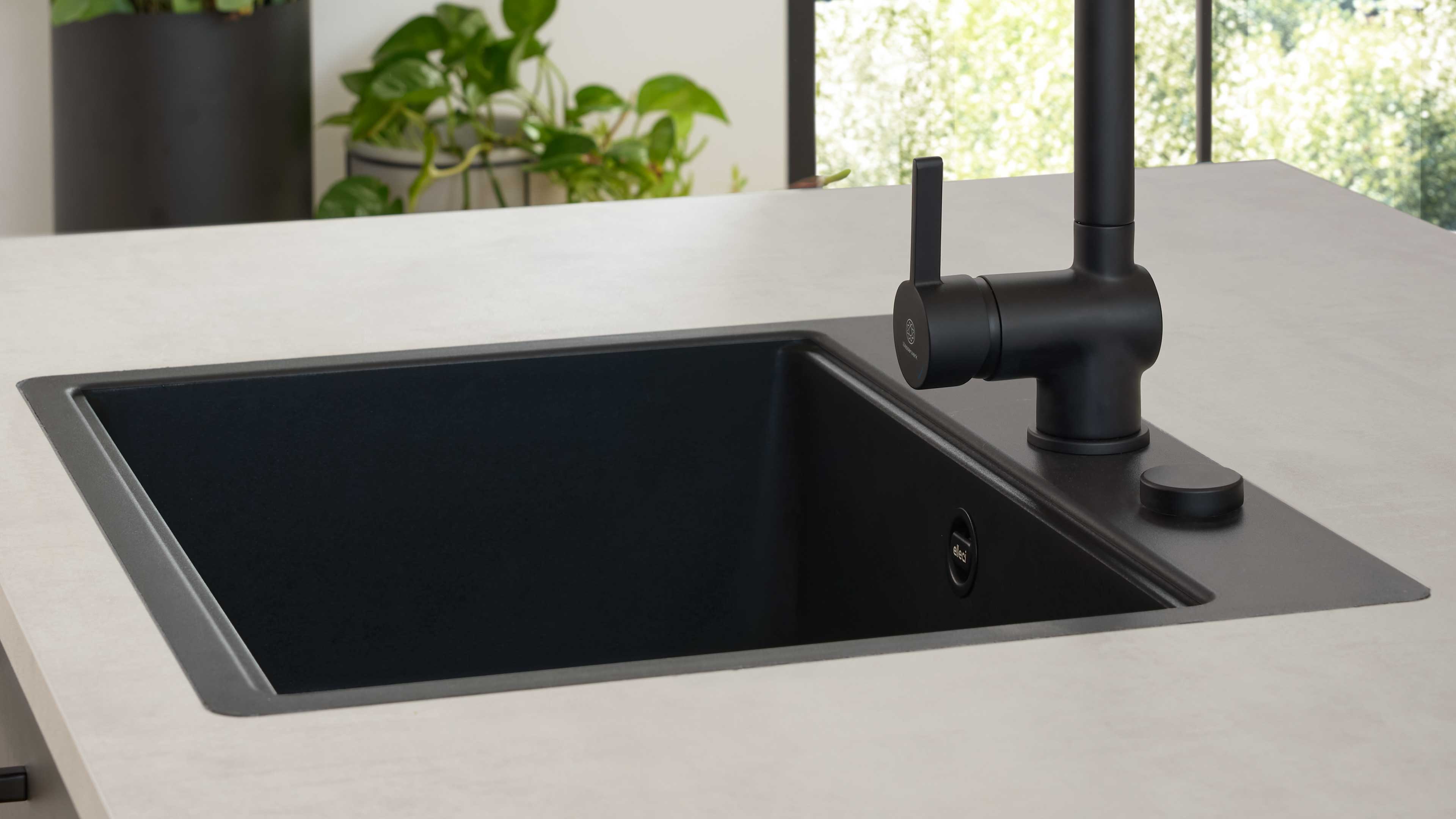 Schwarze eckige Küchenspüle mit breitem Rand in heller Arbeitsfläche, matte schwarze Armatur rechts und Topfpflanze im Hintergrund.