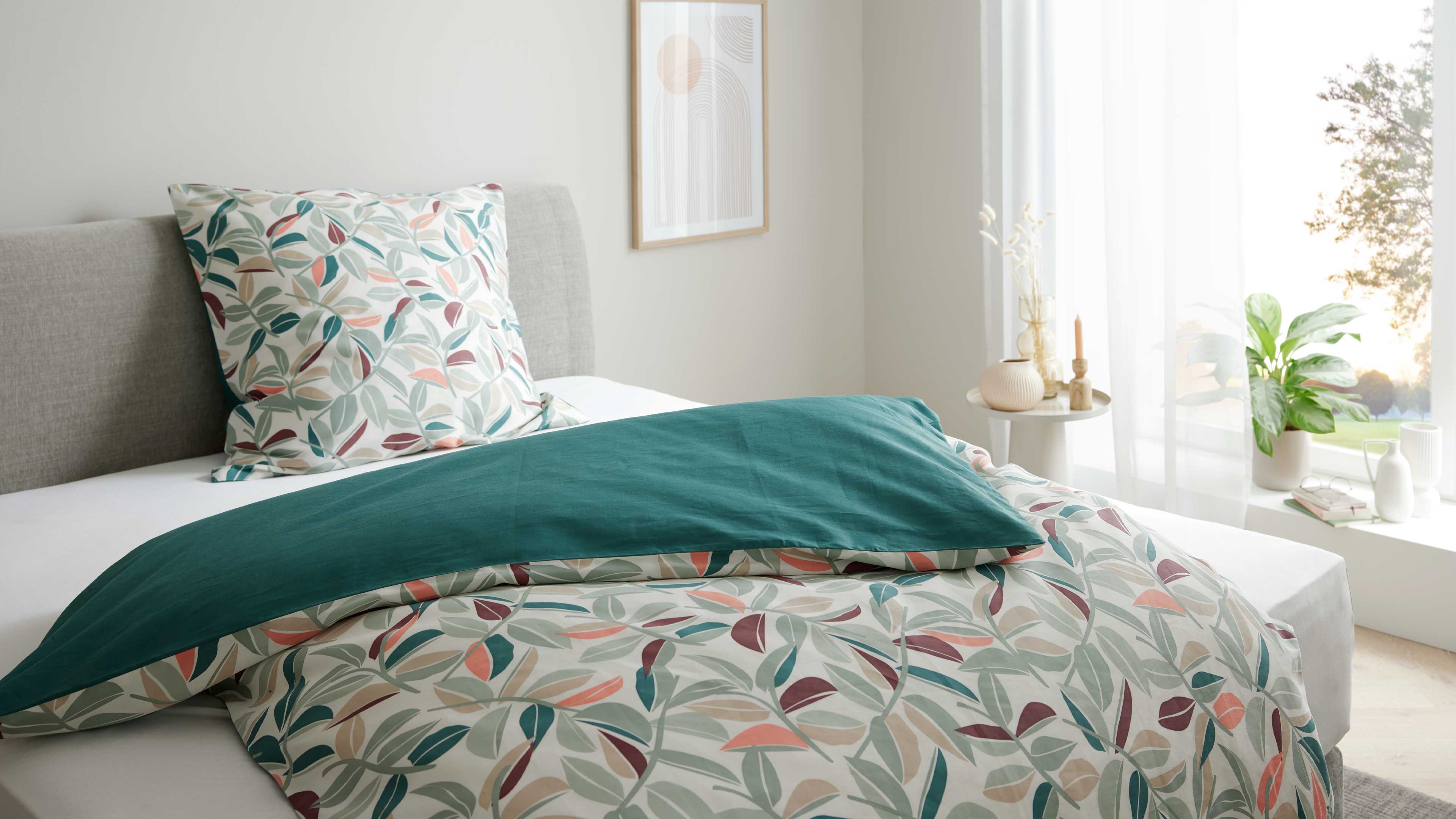 Bett mit grauem Polsterkopfteil, gemustertem Bettbezug mit Blattmotiv und grünem Überwurf, Beistelltisch mit Vasen und Kerze am Fenster.