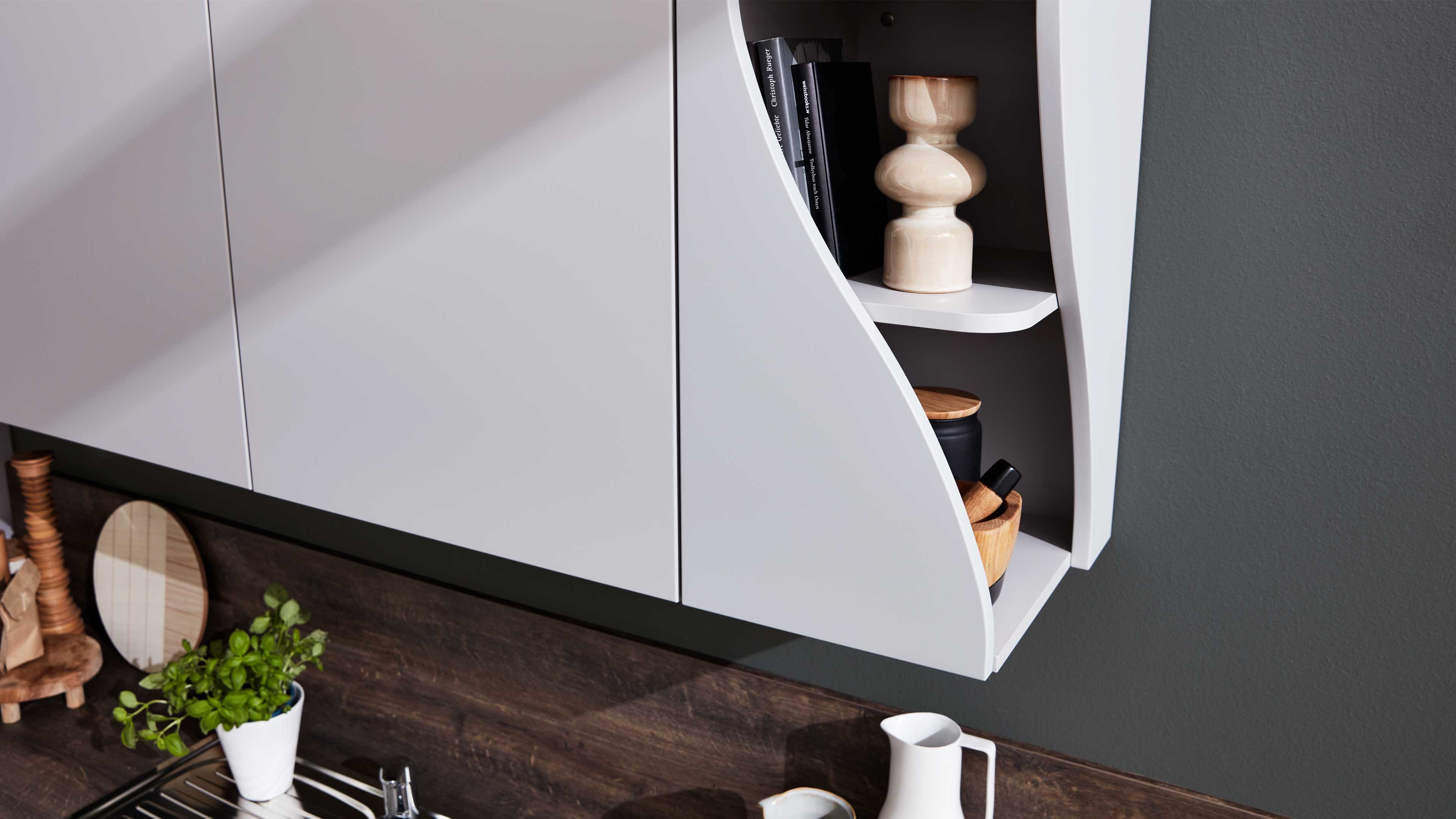 Weißer Hängeschrank mit geschwungener offener Seitenablage, darin Vase, Bücher und Holzschalen, darunter Arbeitsplatte mit Kräutertopf.