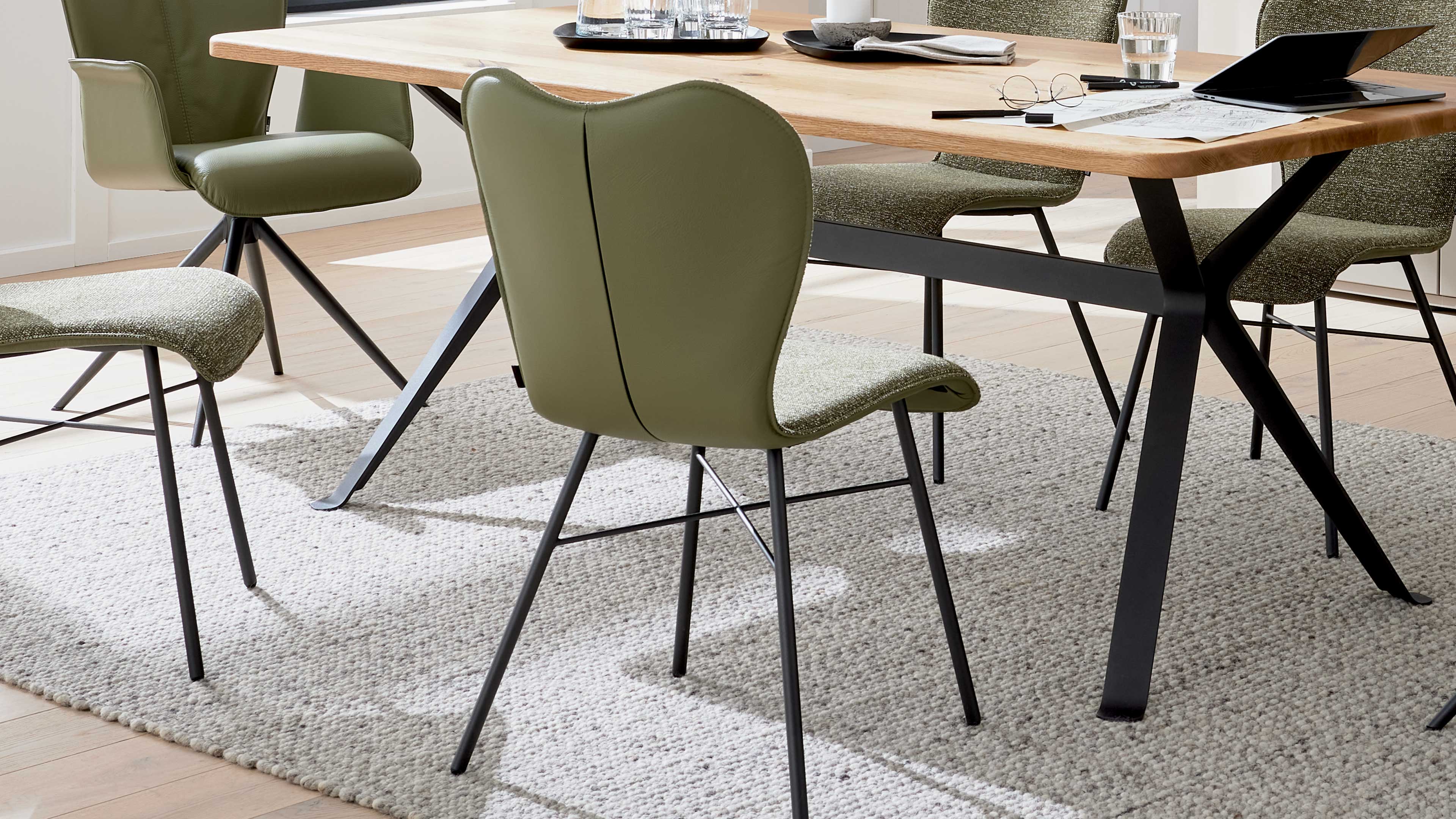 Vordergrund ein olivgrüner Stuhl mit schwarzem Metallgestell vor einem Holztisch mit X-förmigen Beinen auf hellem Teppich.