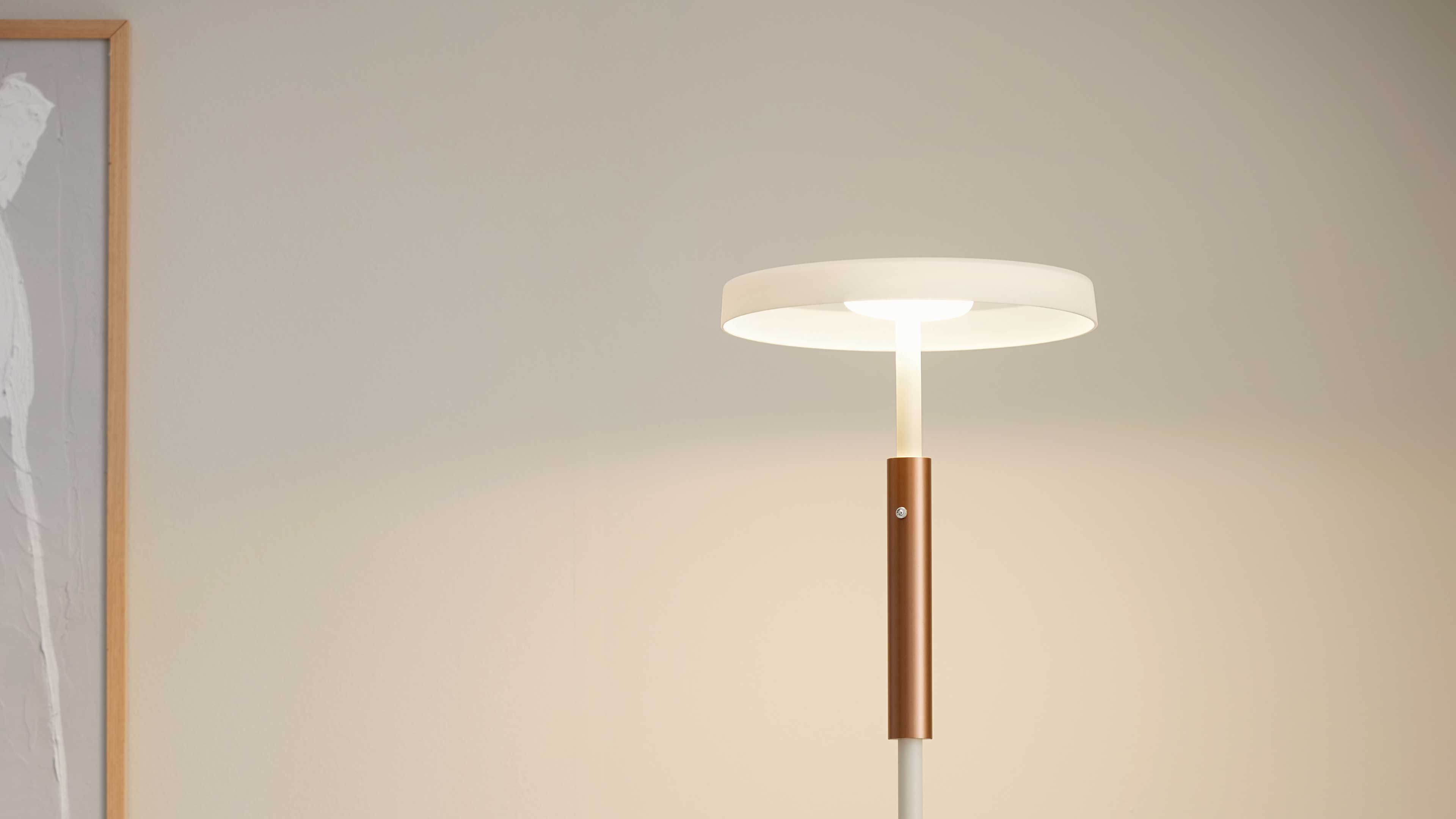 Standlampe mit flachem weißen Rundschirm, schmalem Stab und kupferfarbenem Zylinderdetail, links teilweiser Holzrahmen an der Wand.