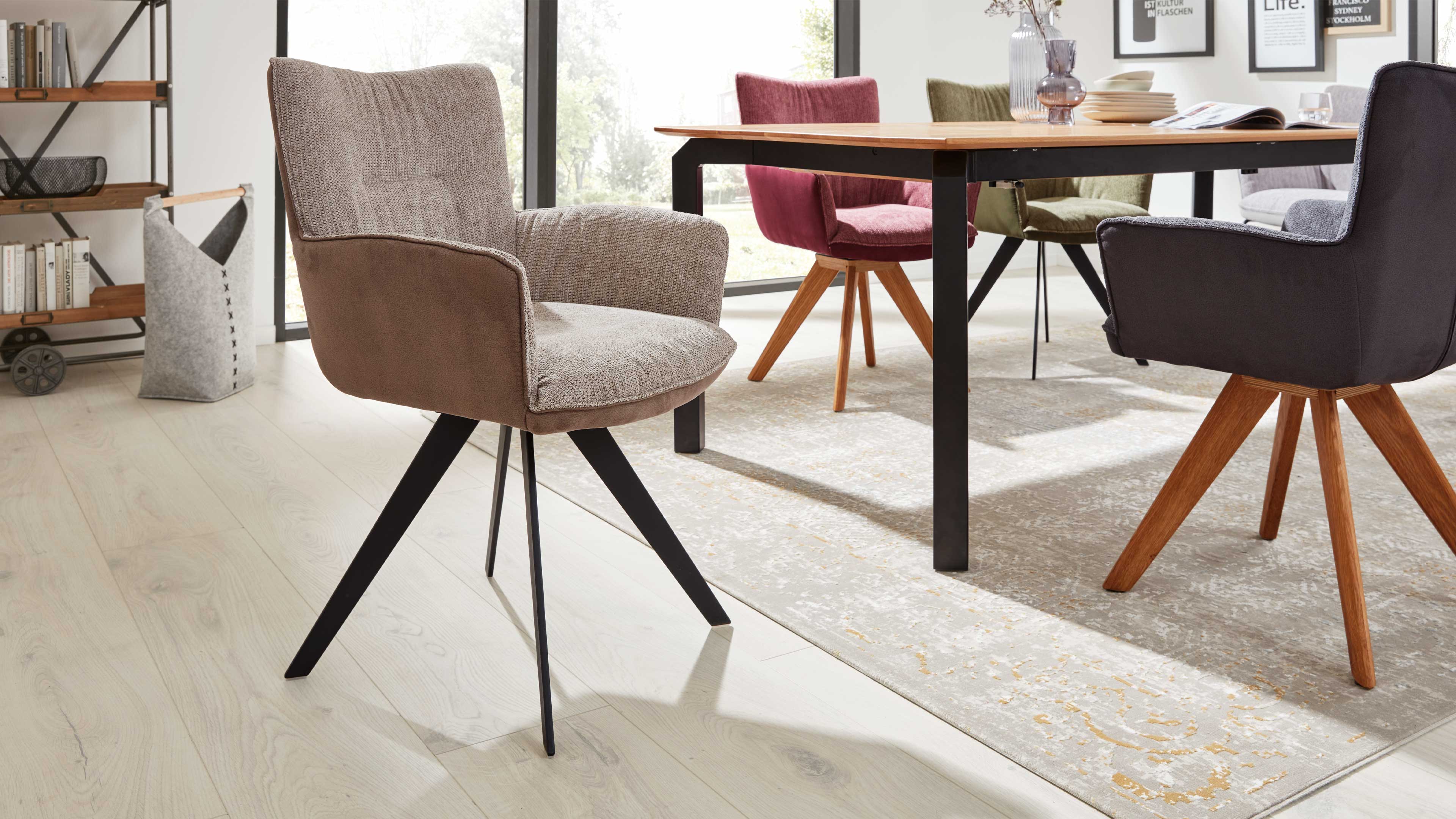 Beiger Polsterstuhl mit geschwungener Rückenlehne und schwarzen schrägen Metallbeinen im Vordergrund, dahinter Esstisch mit bunten Stühlen.