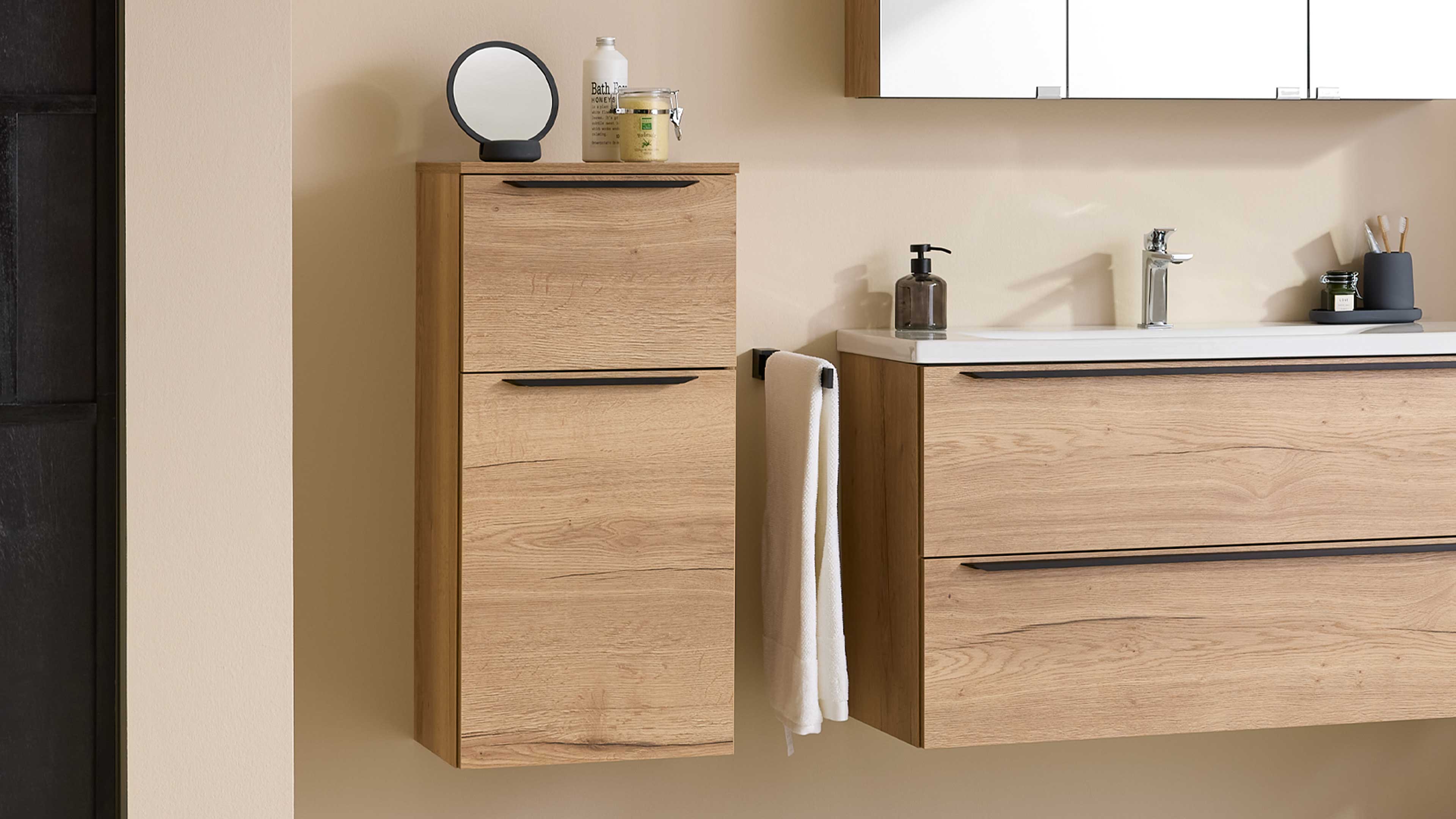 Wandhängender Hochschrank in hellem Holzton mit schwarzen Griffen, daneben Waschbeckenunterschrank, Handtuch und Aufsatzspiegel.