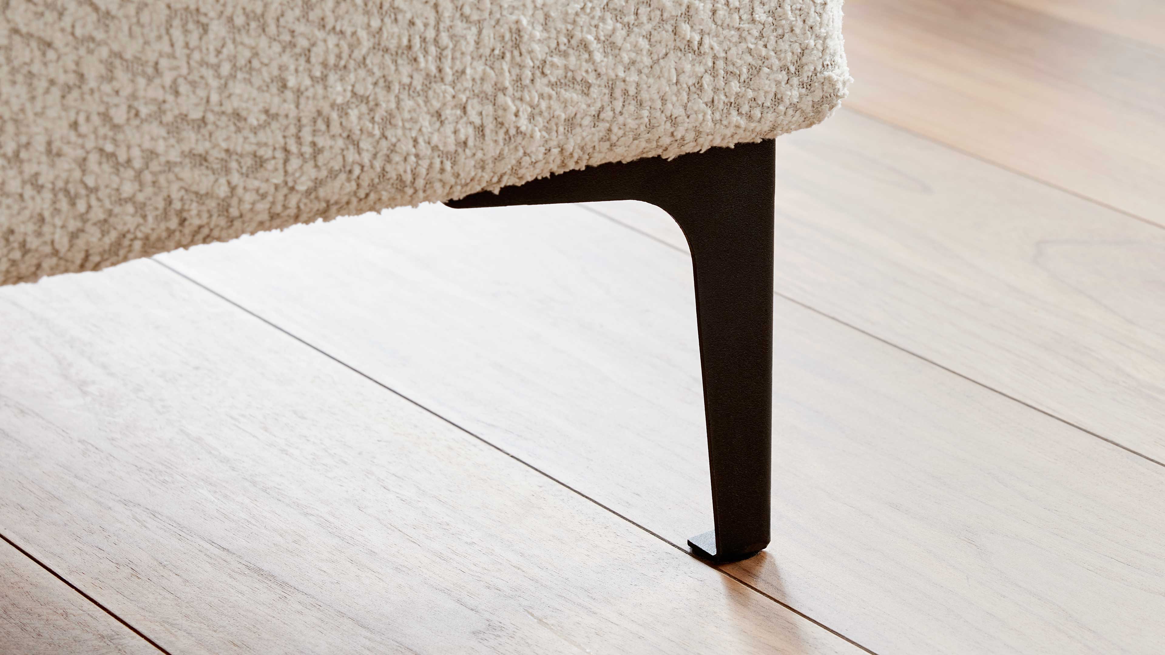 Schwarzes, schlankes Metallbein eines gepolsterten Möbels unter cremefarbenem Bouclé-Stoff, stehend auf hellem Holzboden.