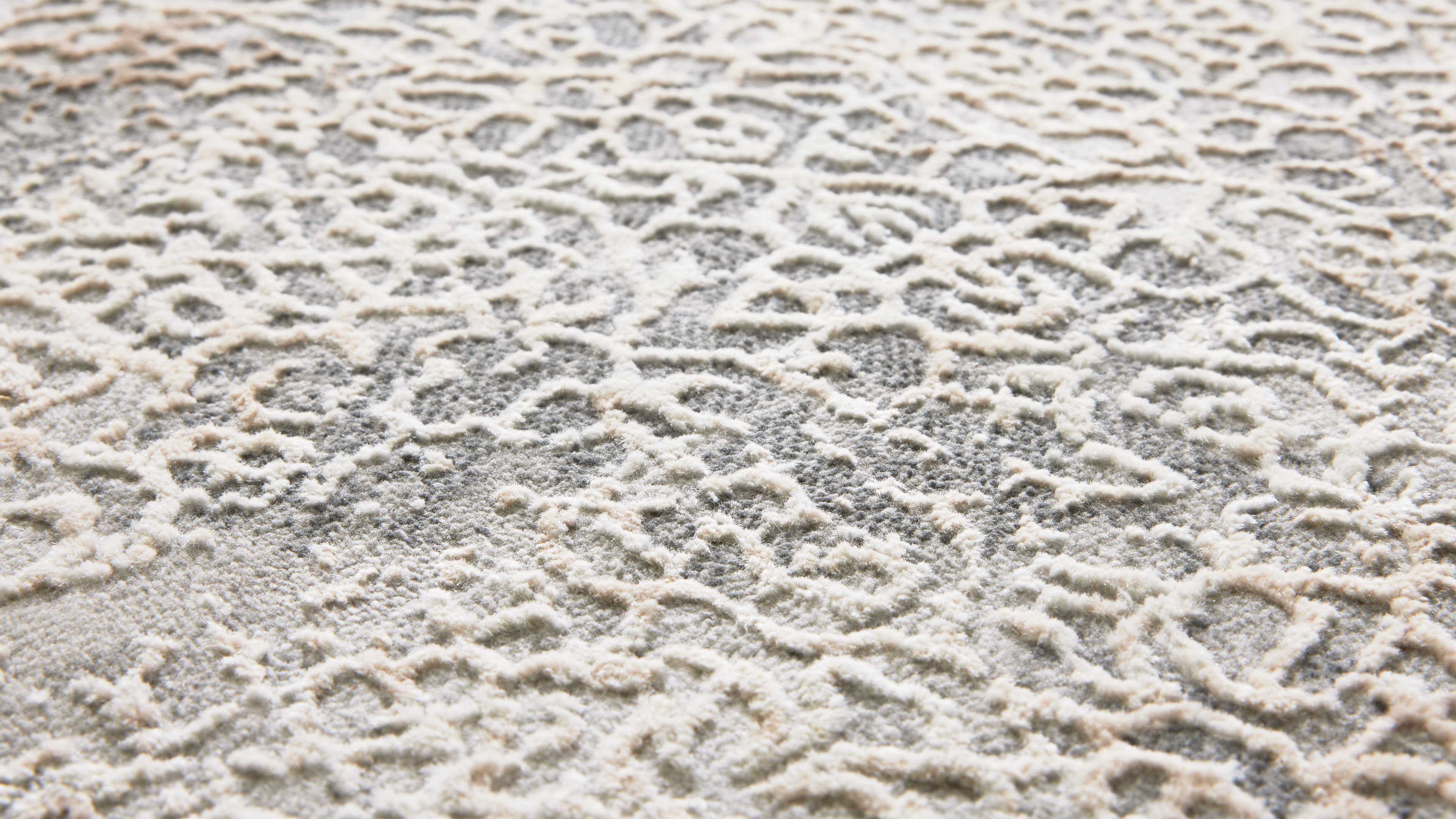 Nahaufnahme eines hellgrauen Teppichs mit cremefarbenen, erhabenen, verschlungenen Schlaufenmustern und texturierter Oberfläche.