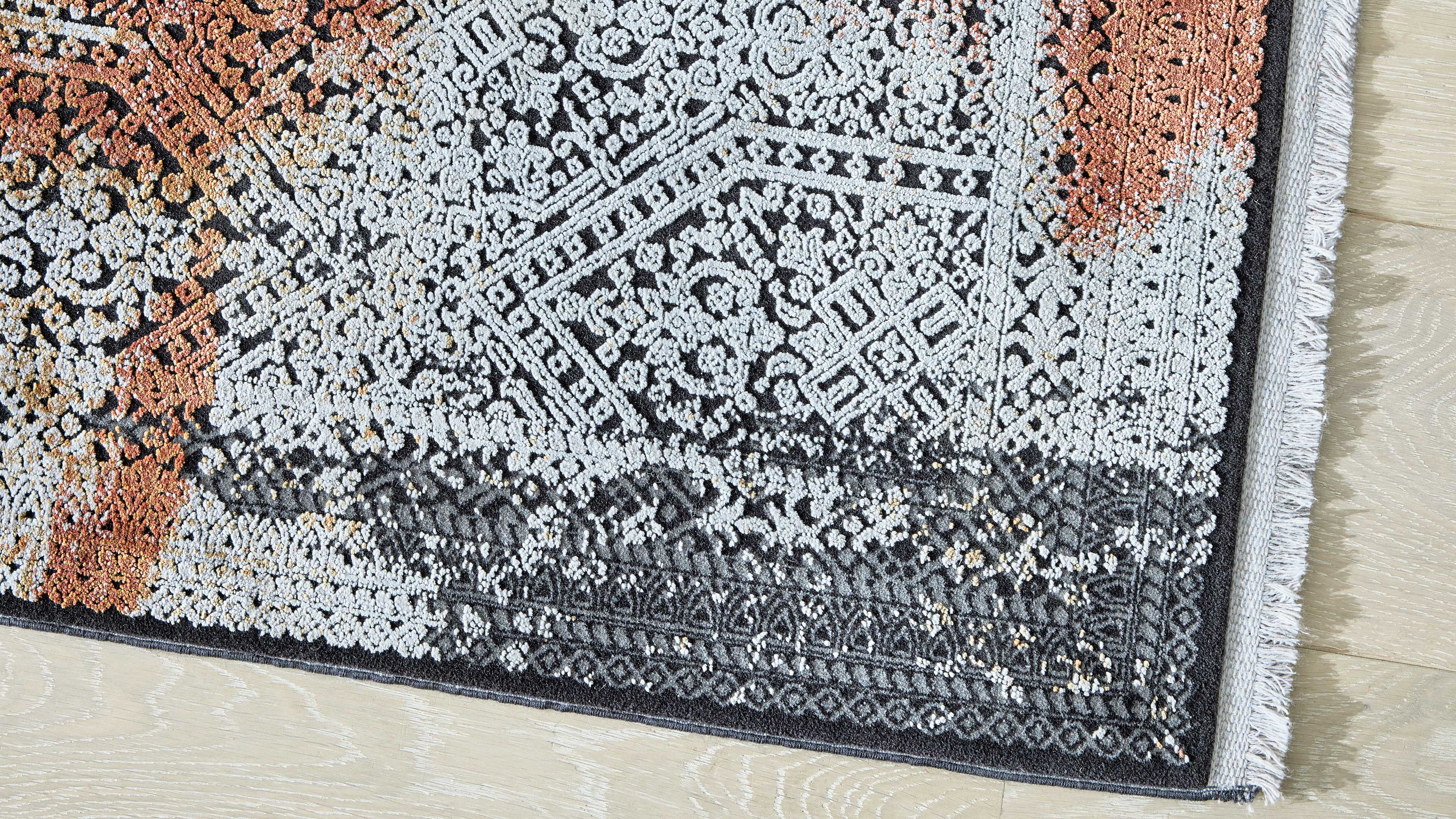 Eckdetail eines gemusterten Teppichs mit Fransen, schwarz-grau-weißem Ornament und rostorange Akzent auf hellem Holzboden.