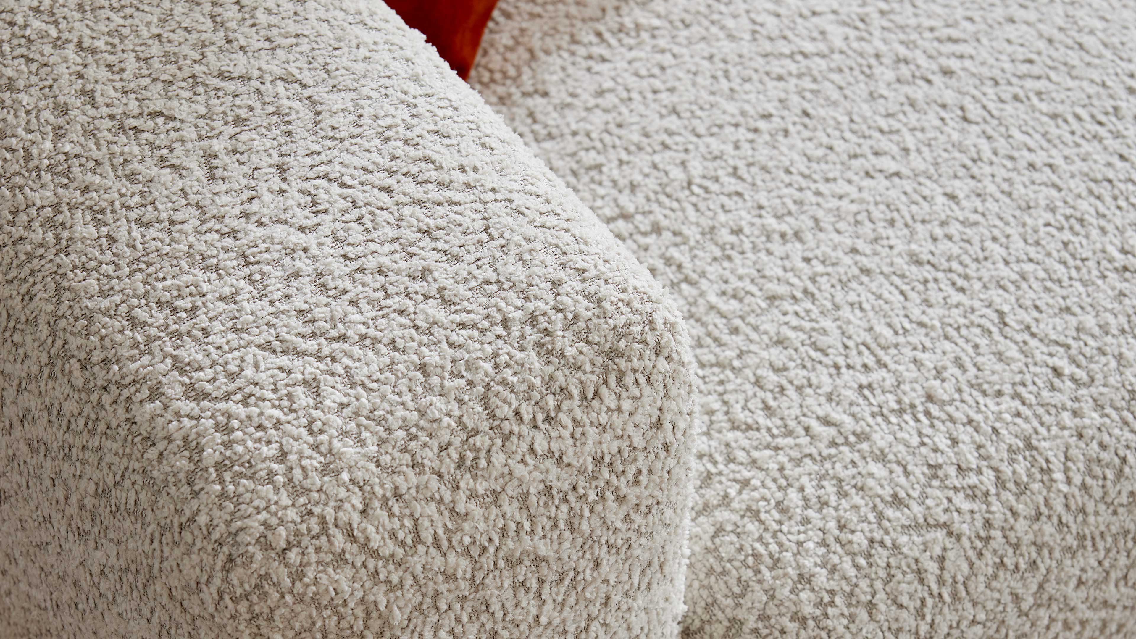 Nahaufnahme einer cremefarbenen Bouclé-Polsterung, die eine abgerundete Armlehne und angrenzende Sitzfläche zeigt; hinten ein rotes Kissen.