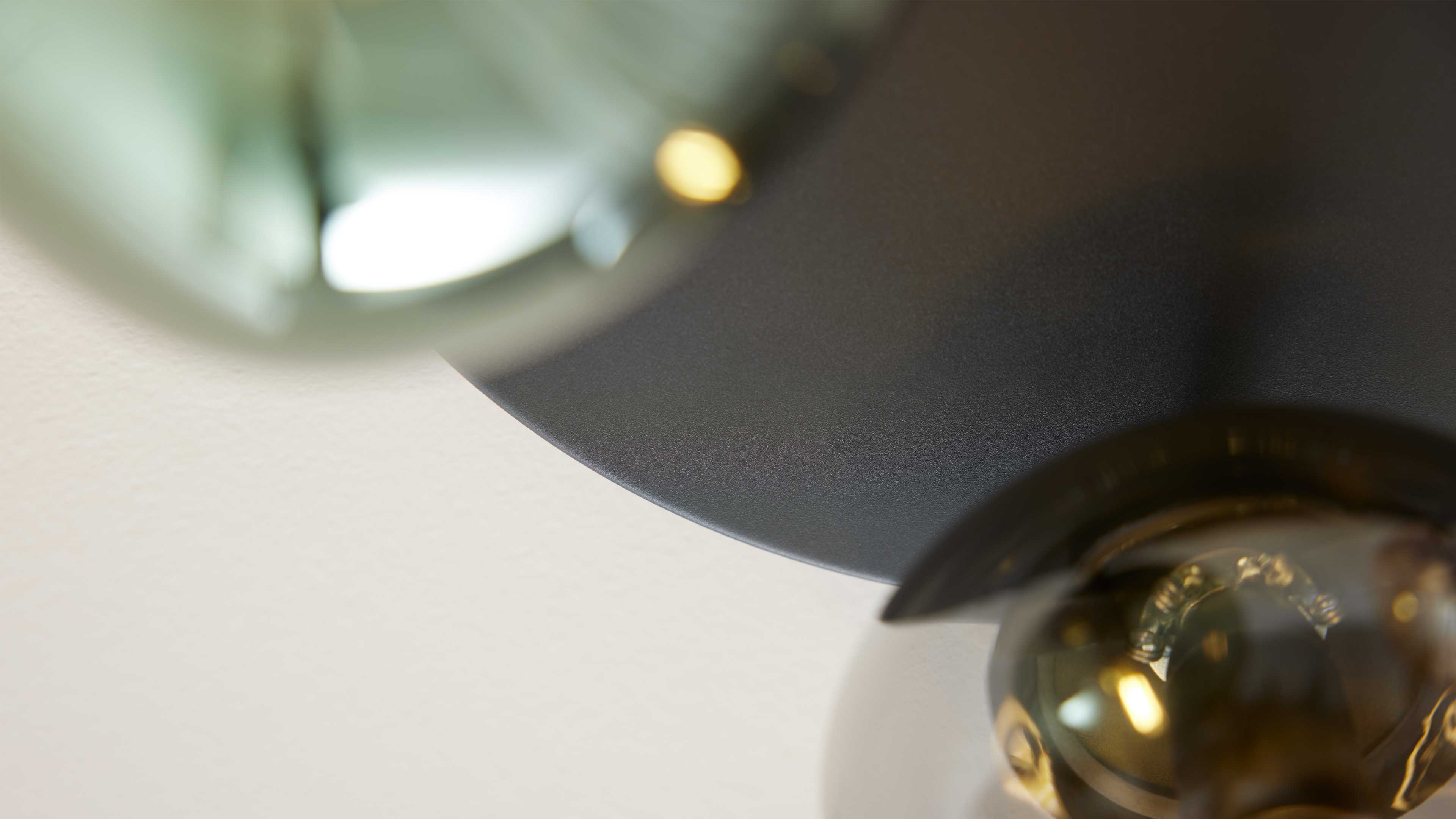 Teil einer Deckenleuchte mit mattem, dunklem Metallschirm und einer runden Glaskugel mit sichtbarem Birnenfilament vor heller Wand.